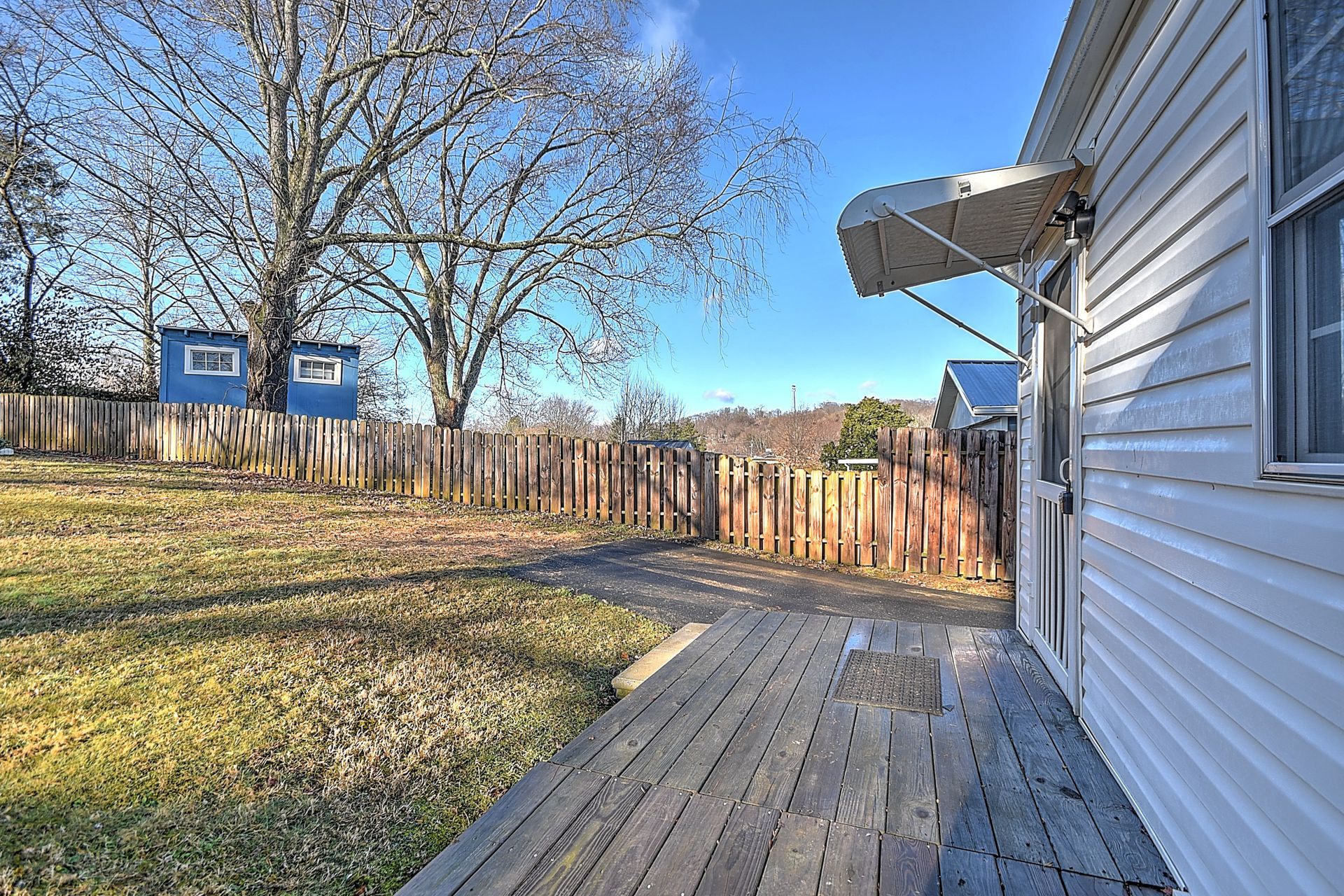 Backyard with wooden deck, fence, small building, and trees against a blue sky.