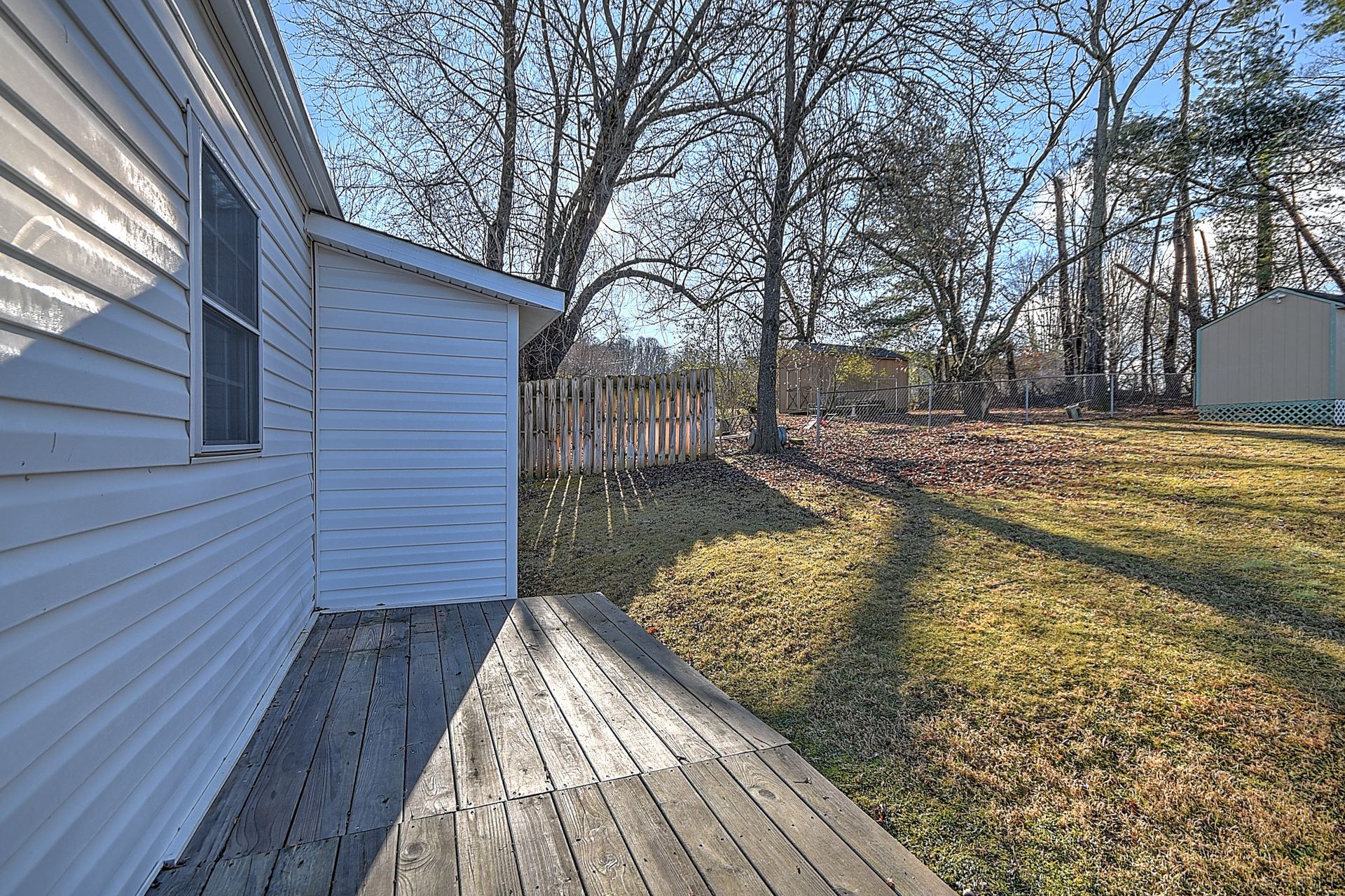 Side view of a house with white siding and a wooden deck overlooking a grassy backyard with trees.