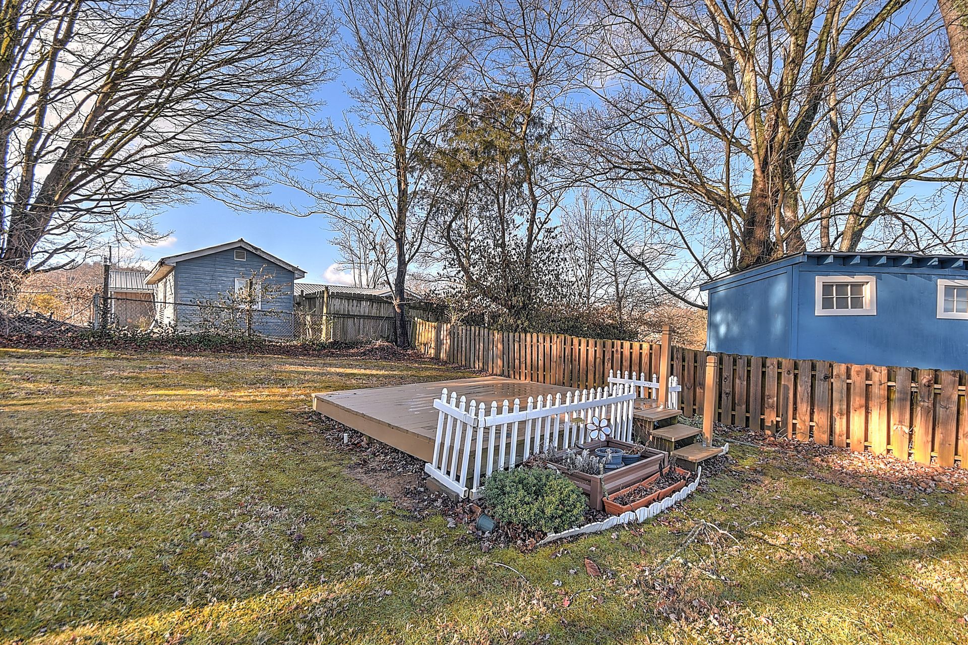 Backyard with deck, picket fence, blue shed, and leafless trees under a blue sky.