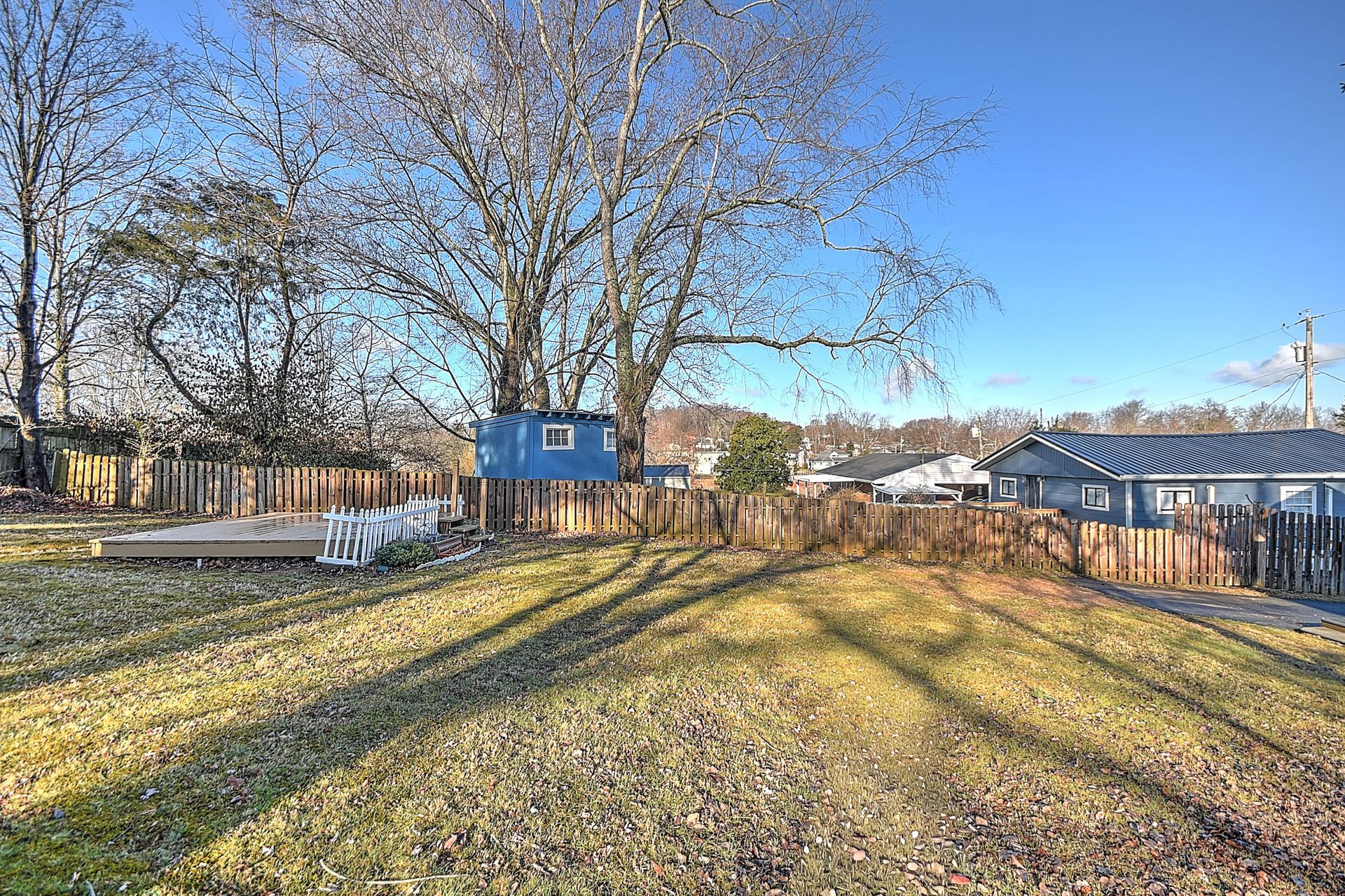 Backyard with brown grass, wooden fence, trees, and blue-painted structures under a clear sky.