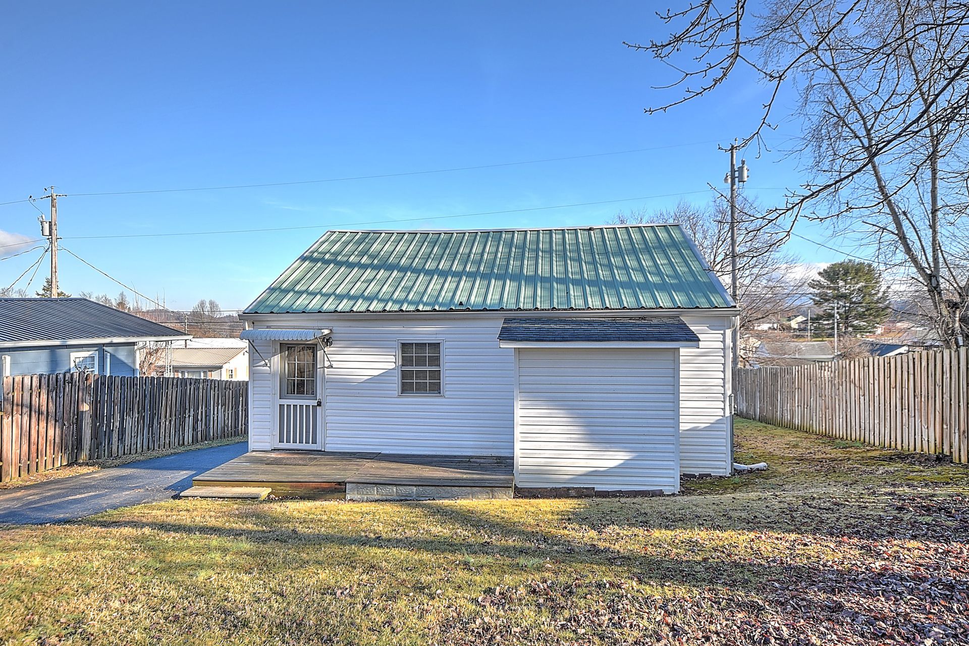 White one-story house with green metal roof, small shed, and brown fence, set on a lawn under a blue sky.