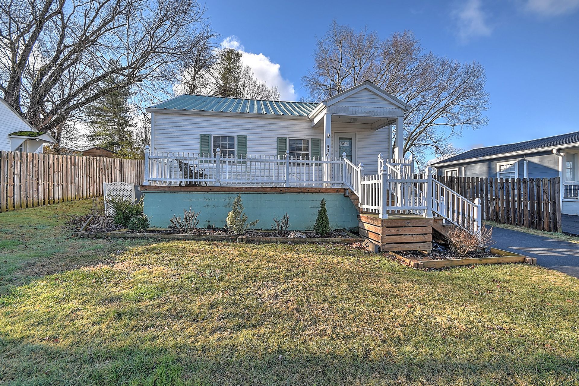 White house with a porch, blue-green foundation, and green metal roof. Brown wooden fence and dry grass in front.