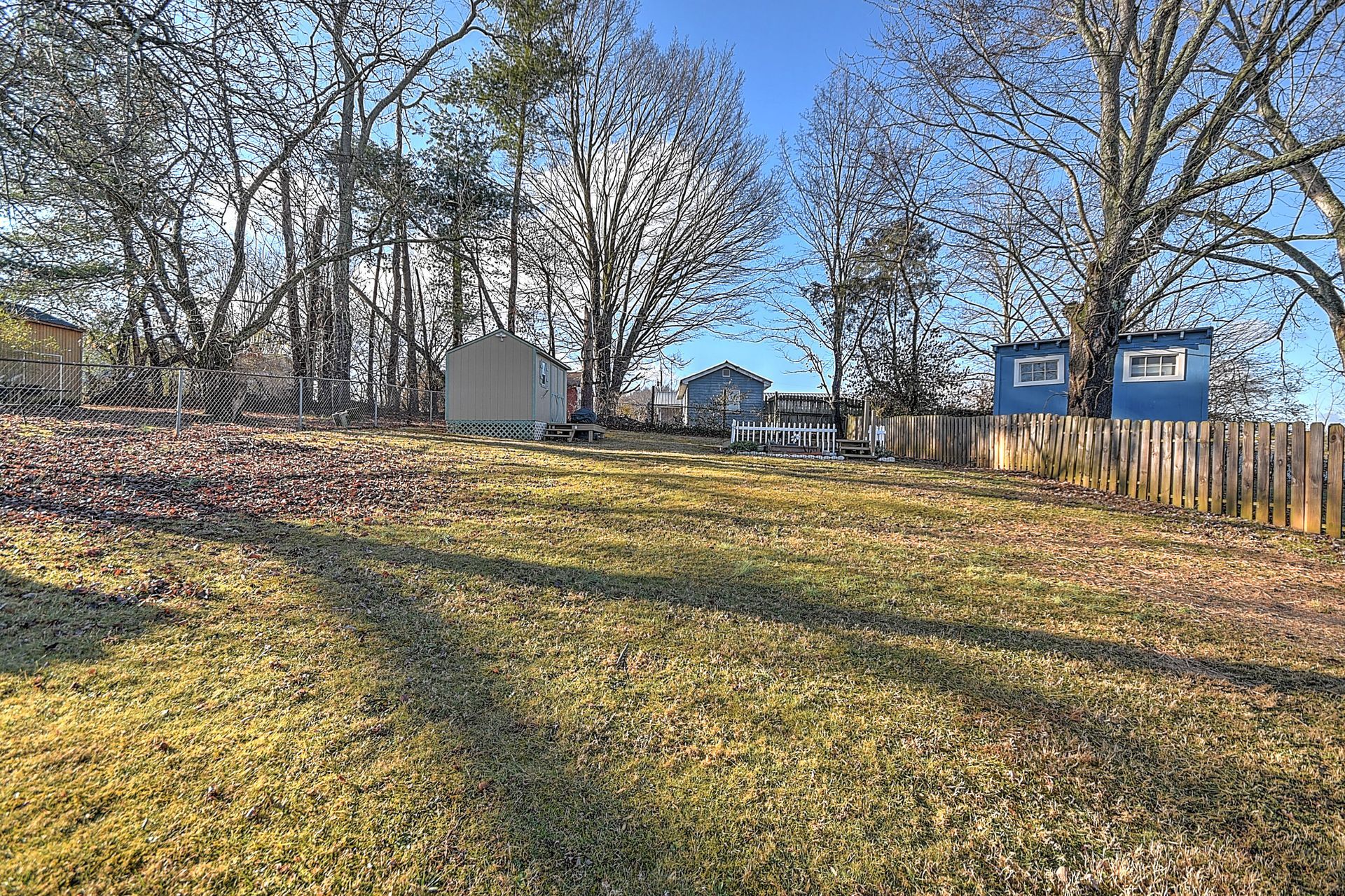 Grassy backyard with shed, small blue building, and trees against a blue sky.