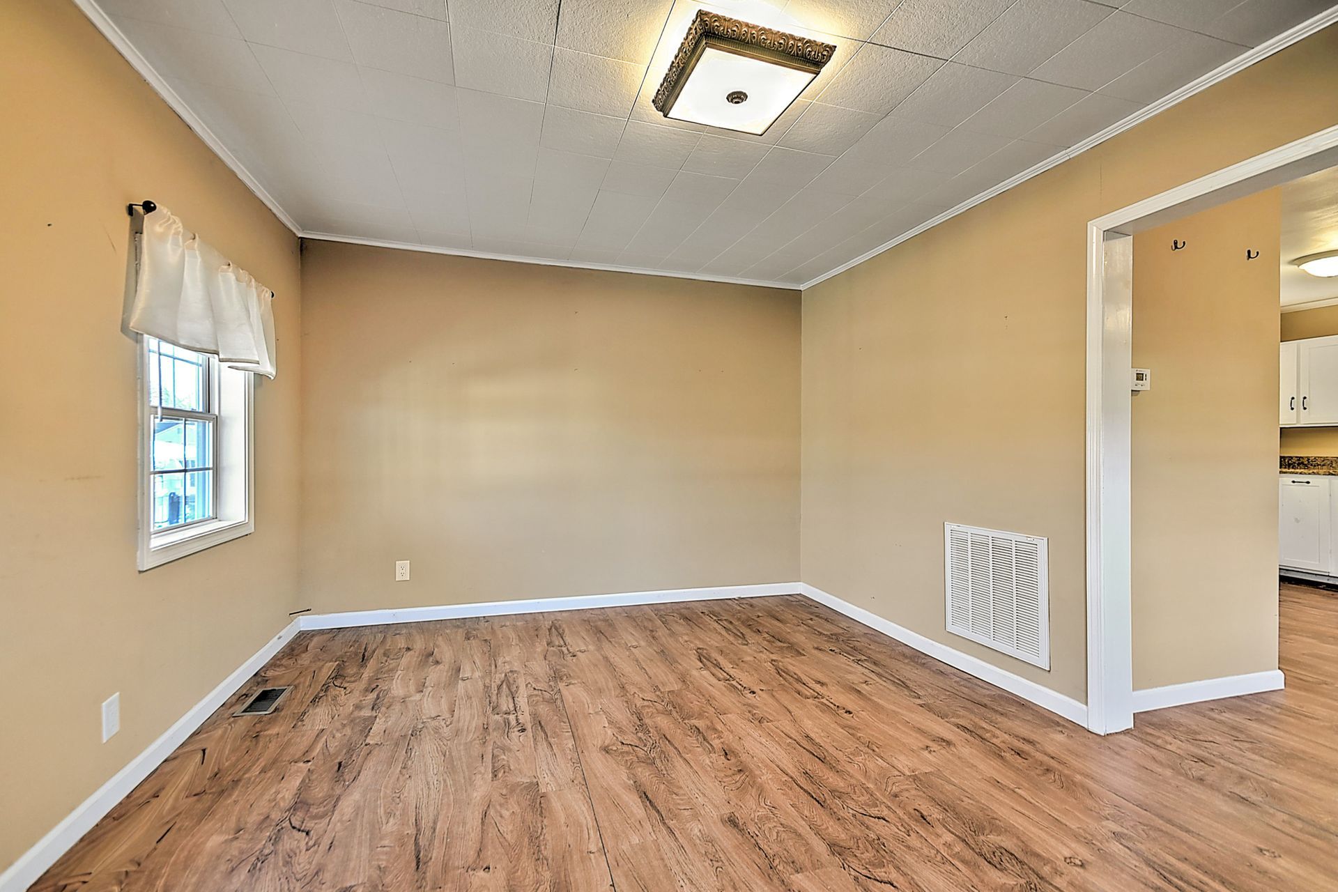 Empty room with tan walls, wood-look floor, window, and doorway to a kitchen.