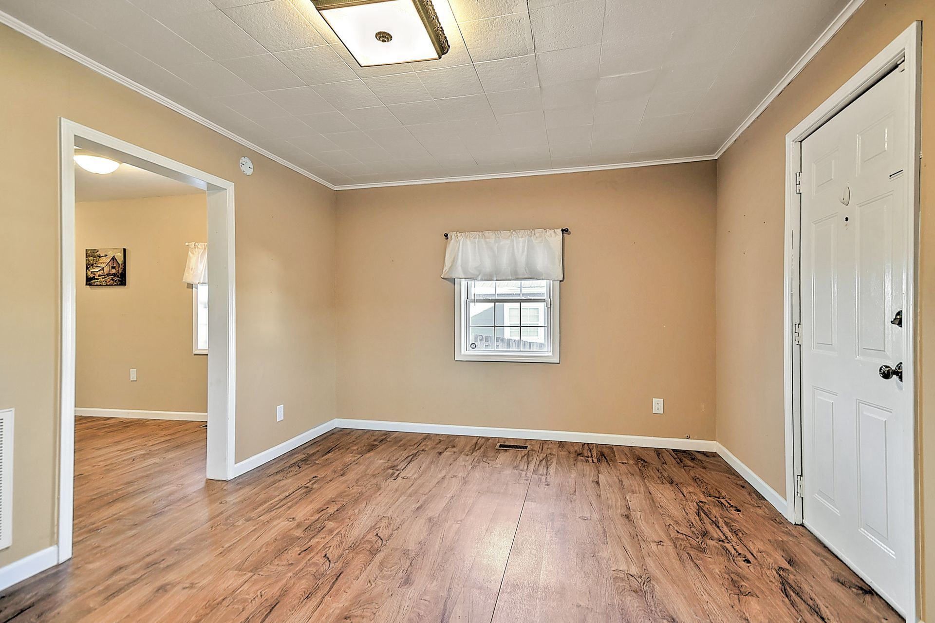 Empty room with wood floors, tan walls, small window, and white door.