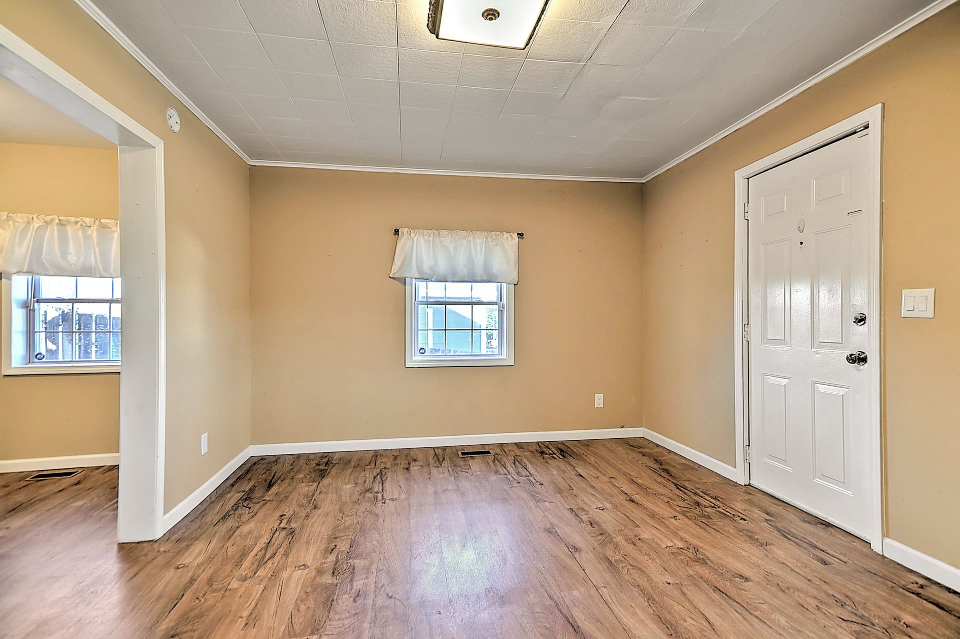 Empty room with beige walls, white door, and wood-look flooring. Window with curtain, and entryway visible.