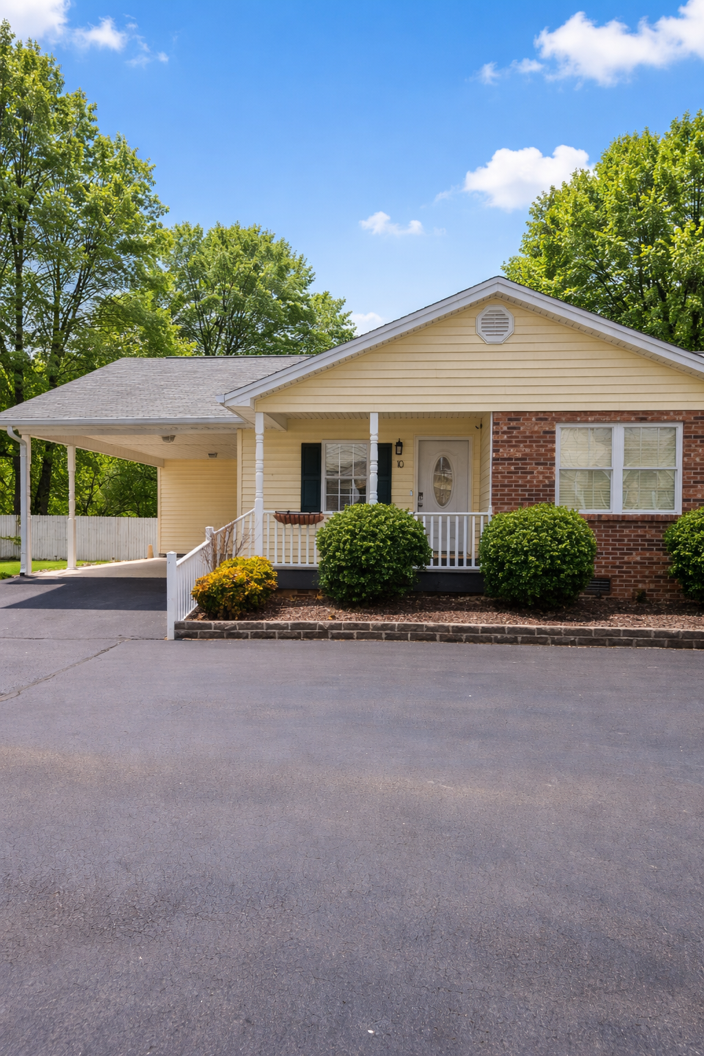 A light yellow one-story house with a brick accent, front porch, and attached carport under a blue sky with lush trees.