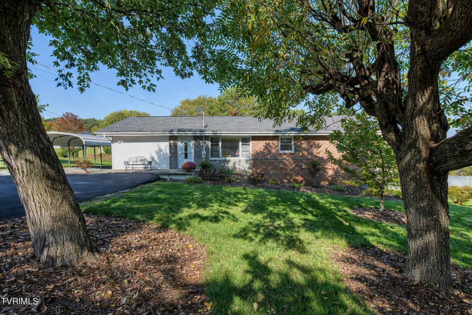 Ranch-style house framed by two trees, set on a green lawn. Asphalt driveway and carport are visible.