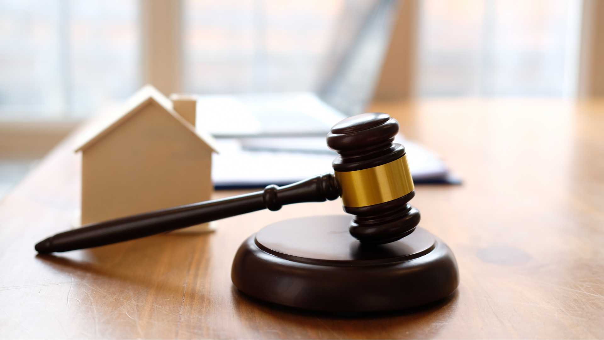 Wooden gavel on stand next to a small house model, laptop, and documents on a table.