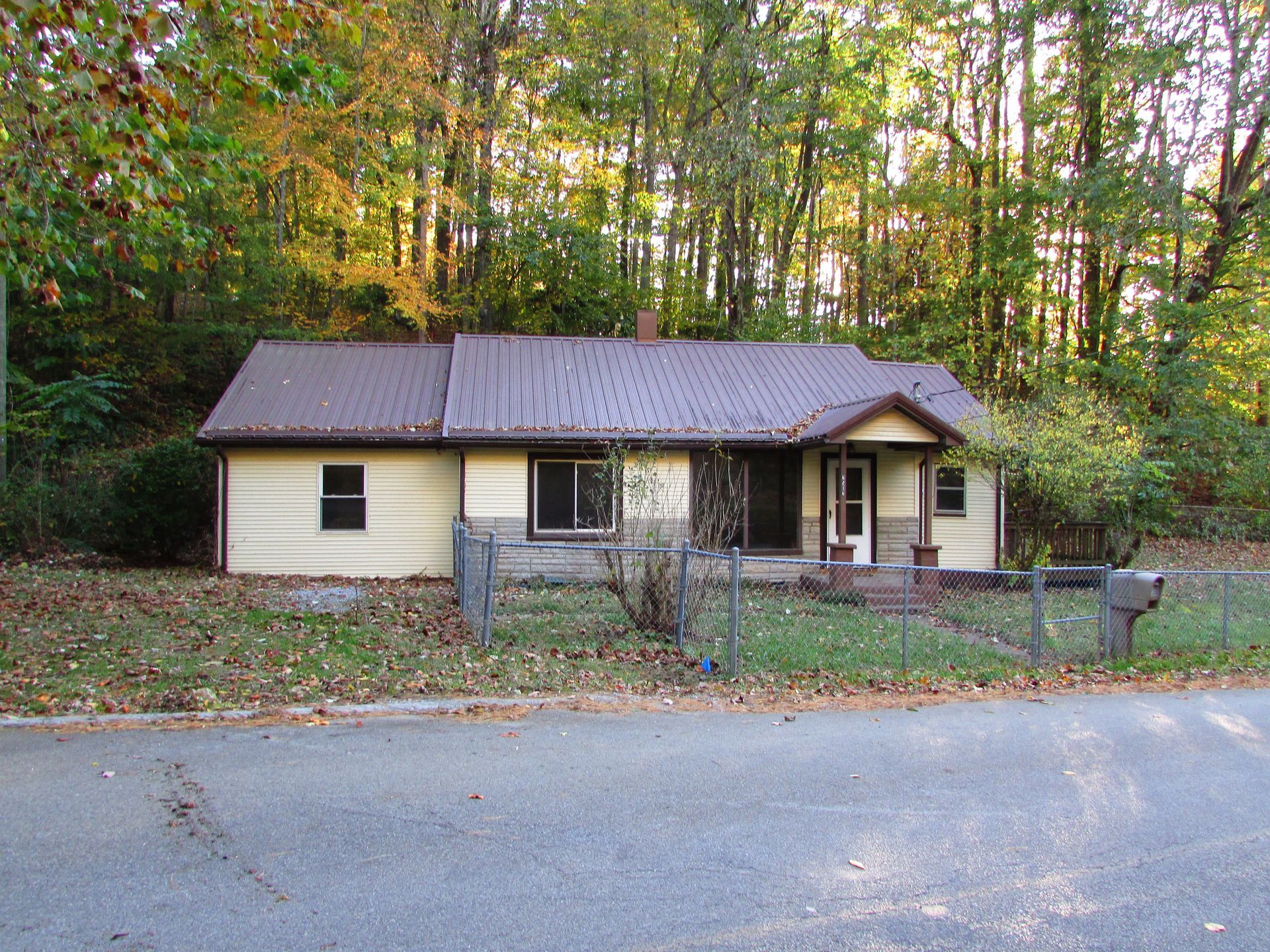 Small, tan house with brown metal roof; front porch; gray fence; trees in background.