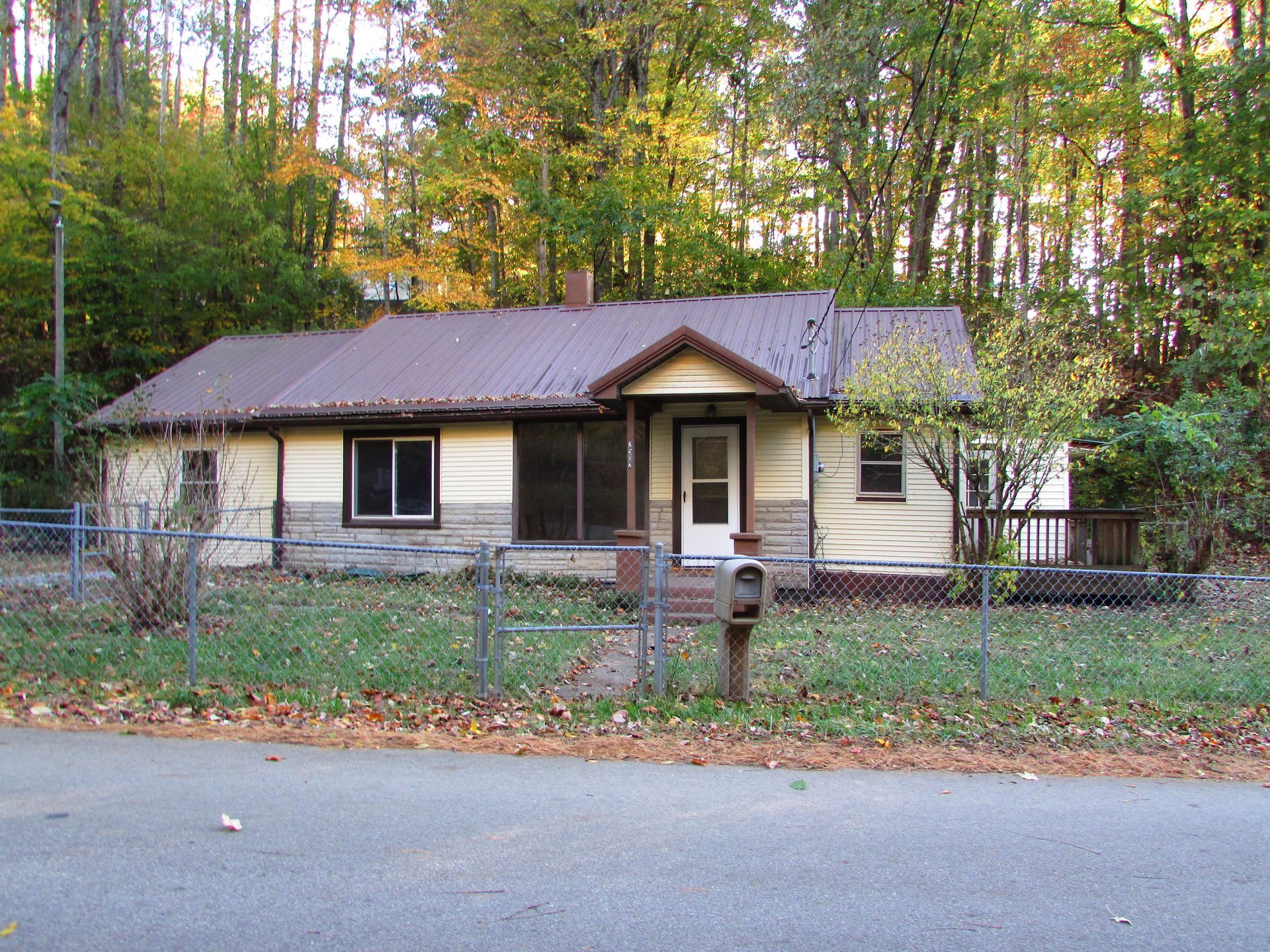 Small, beige house with a brown metal roof. Front porch, fenced yard, surrounded by trees.