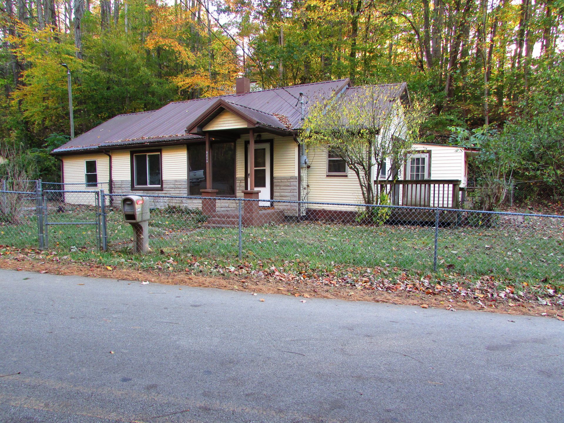 A single-story house with a porch and detached deck, set in front of a forest with autumn leaves.