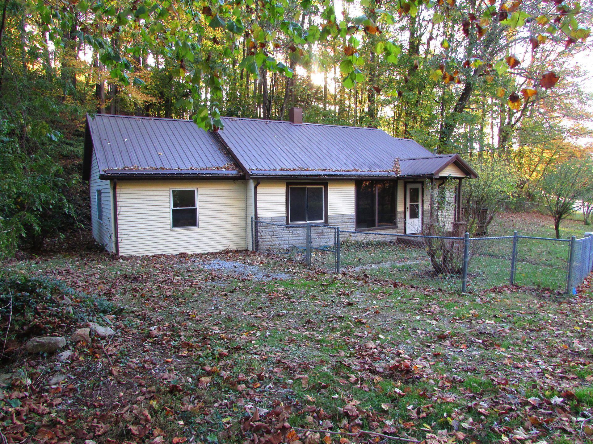 Small beige house with brown roof and chain-link fence on a grassy lot with fall leaves.