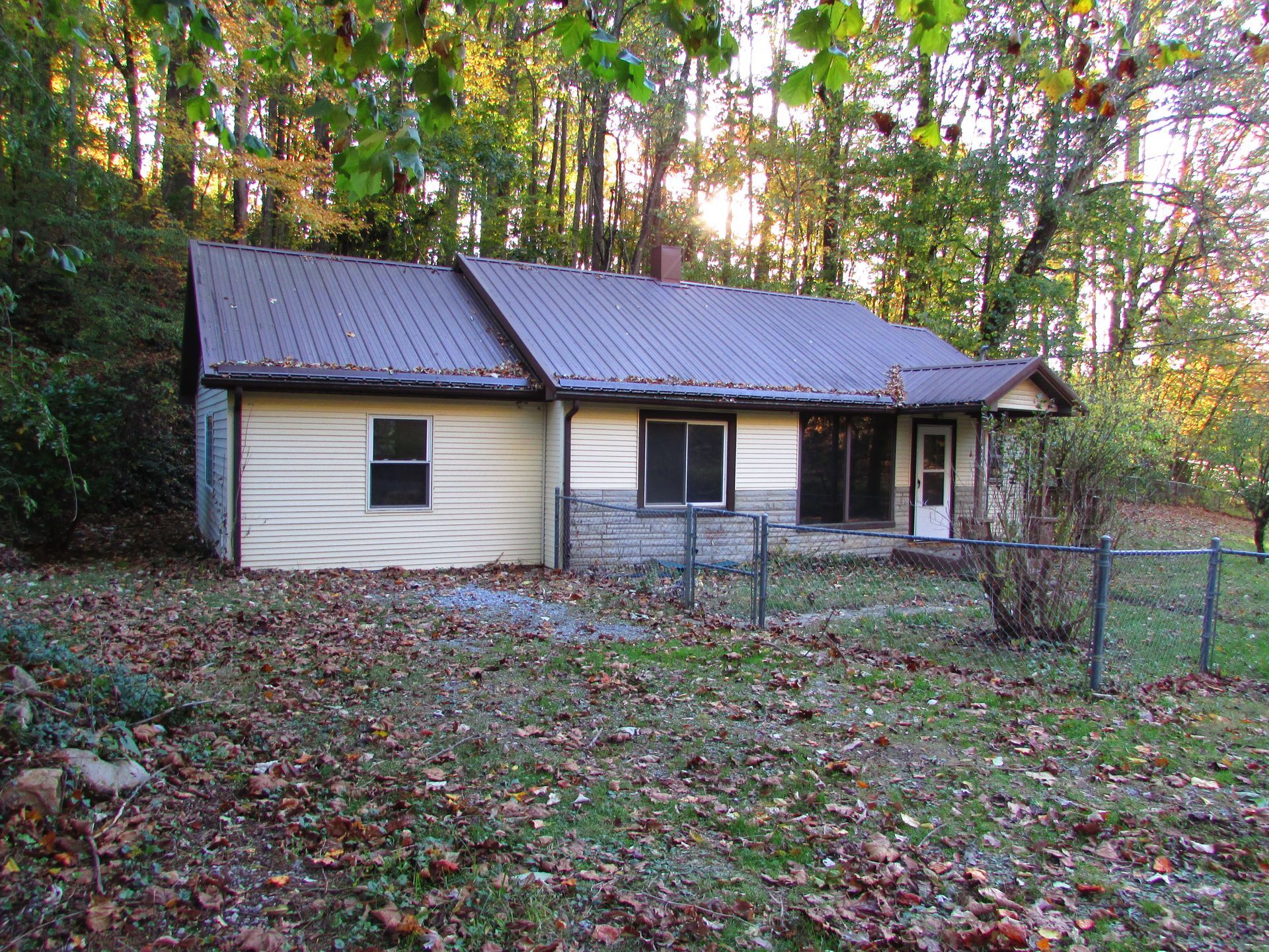 Small beige house with brown metal roof, set in a wooded area, surrounded by a chain-link fence.
