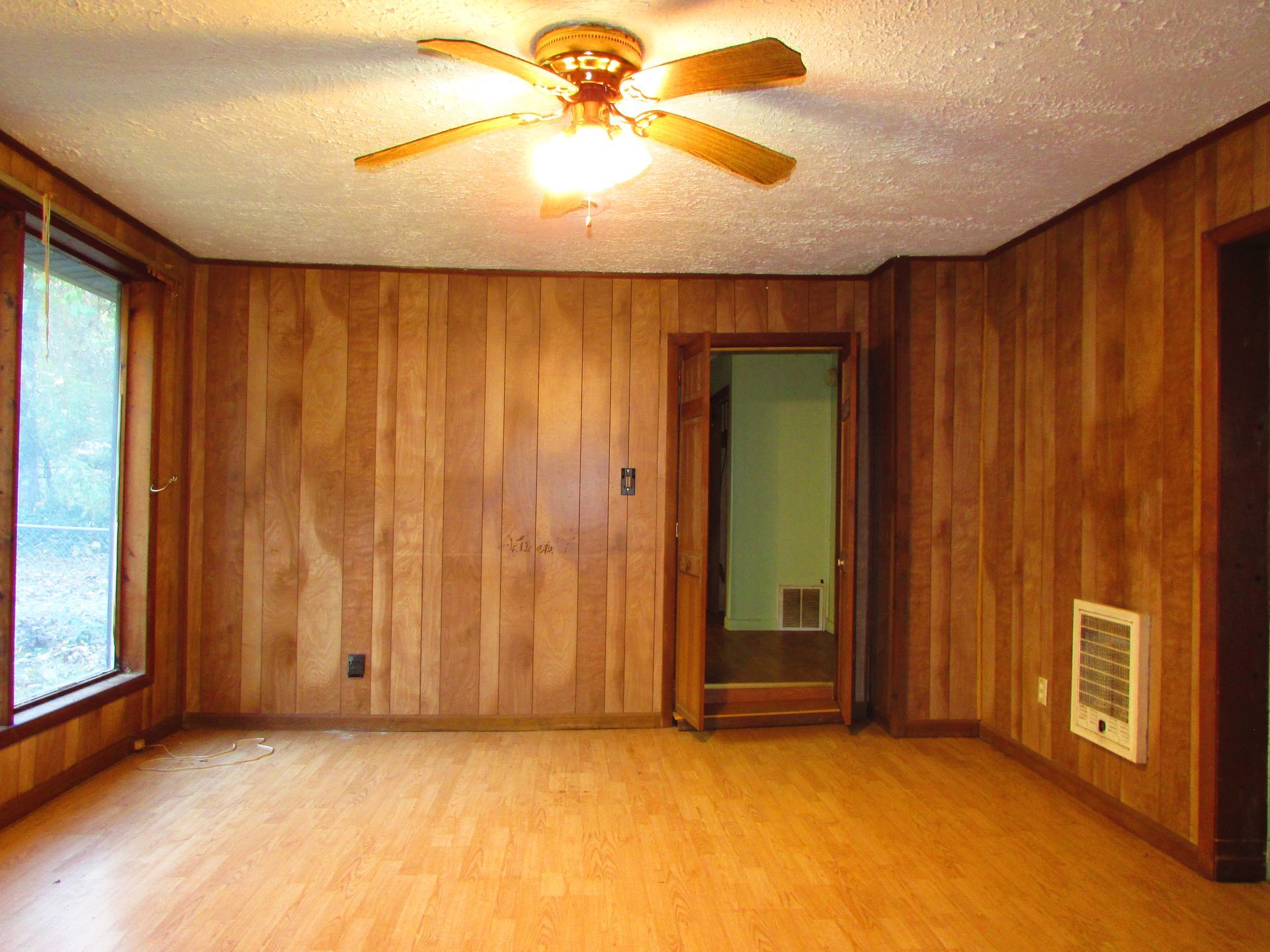 Empty room with wood paneling on walls, door, window, ceiling fan, and heater.