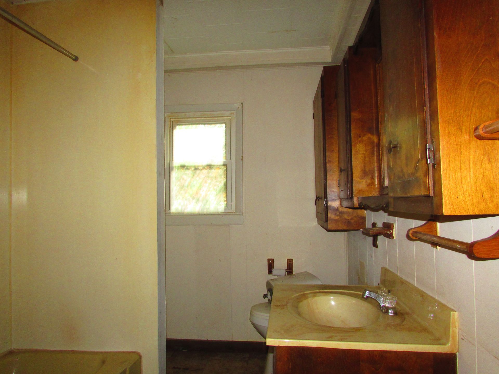 Small, old bathroom with stained cabinets, a sink, a toilet, and a window.