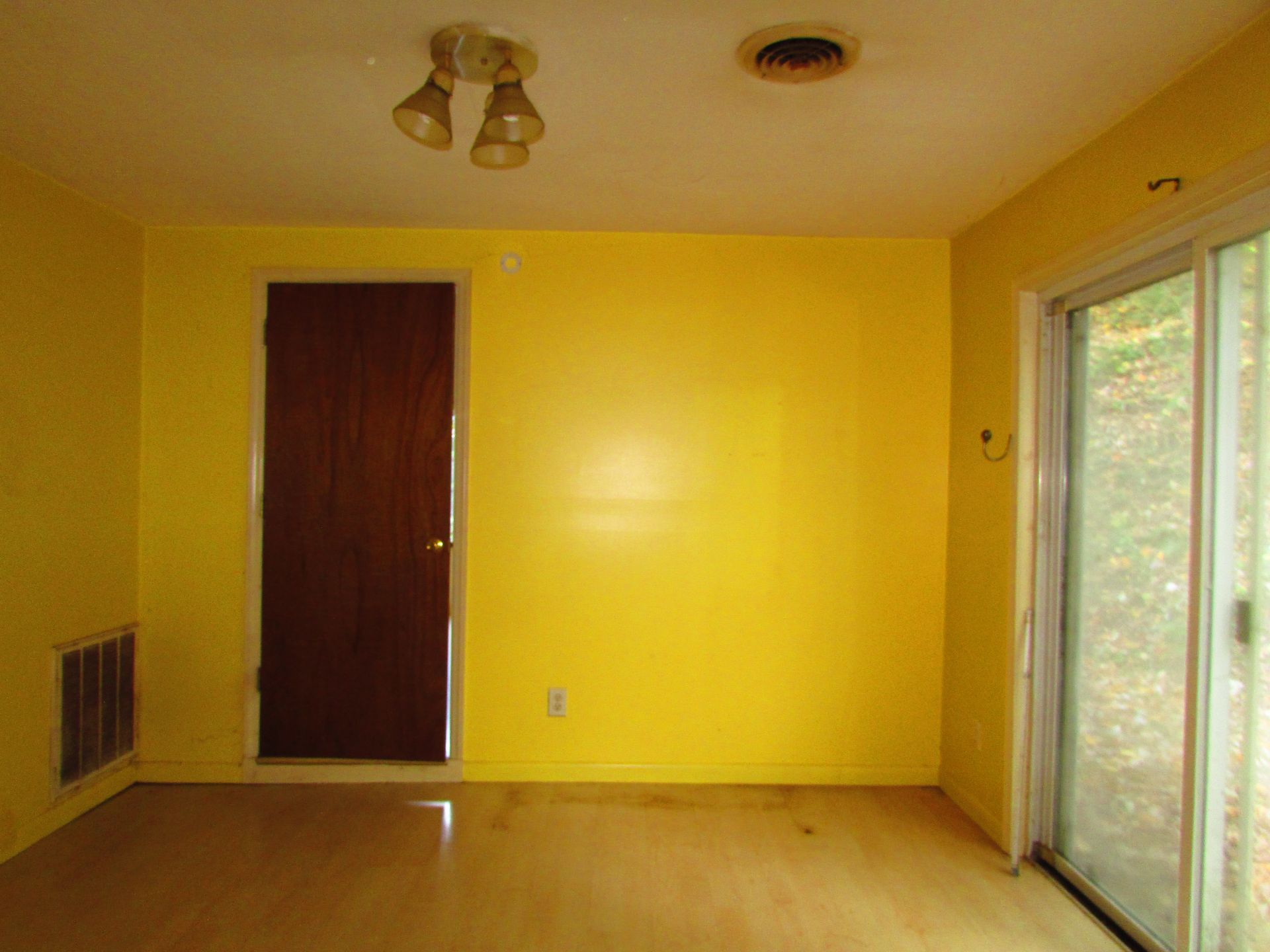 Yellow-walled empty room with a brown door, sliding glass door, and a three-bulb ceiling light.