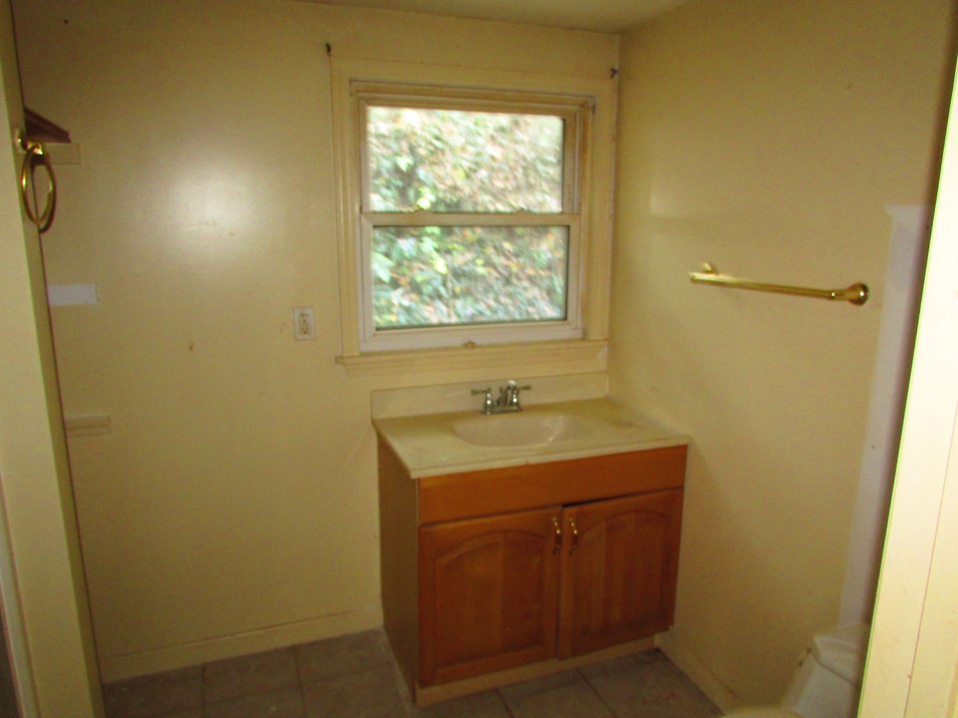 Small bathroom with wood vanity, window, and gold towel rack. Pale yellow walls.