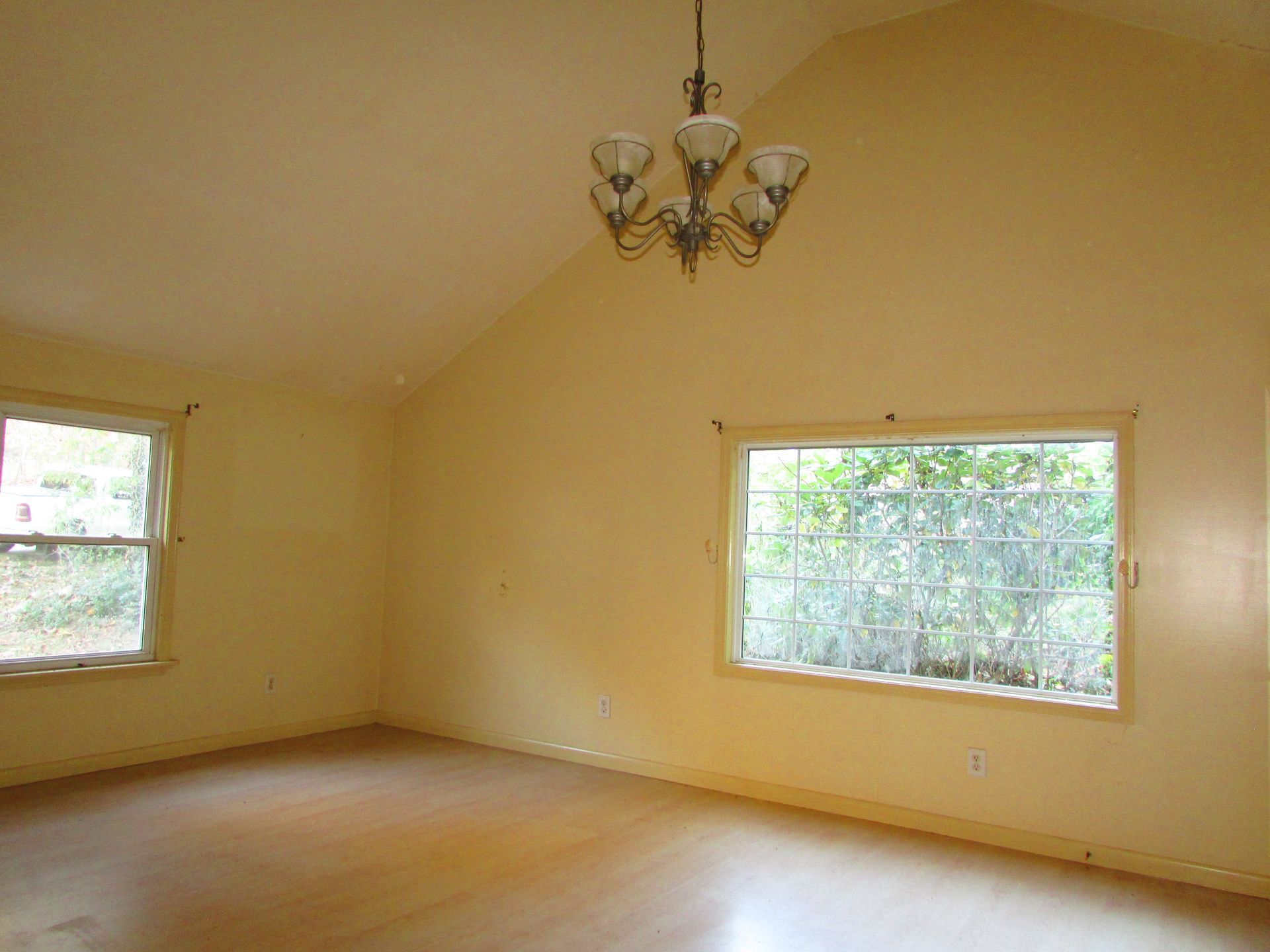 Empty room with pale yellow walls, wooden floor, and a chandelier; two windows.