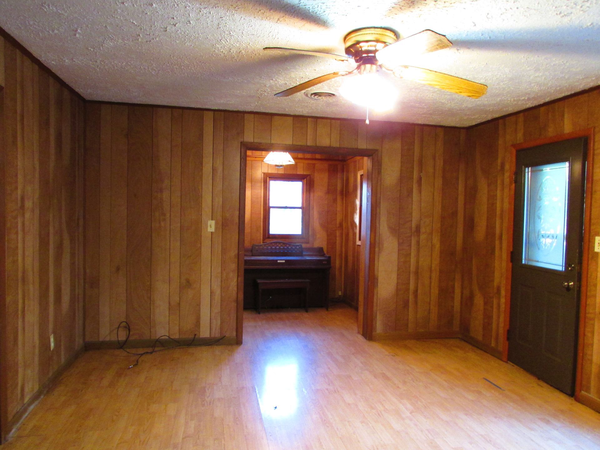 Empty room with wood paneling walls and flooring, ceiling fan, and door.  A doorway leads to another room.