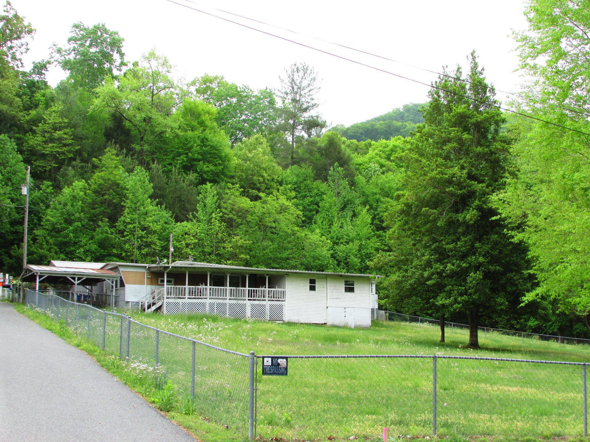 Rural white house beside a road, fenced grassy yard, and dense green trees on a hillside