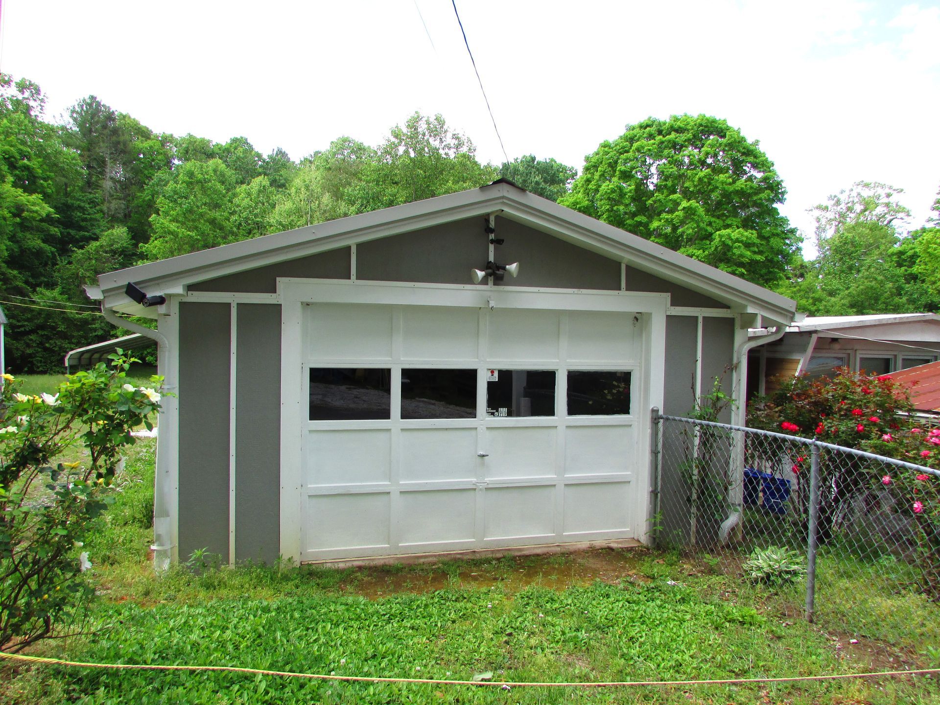 Small gray garage with white door in a grassy yard, surrounded by trees and a chain-link fence.