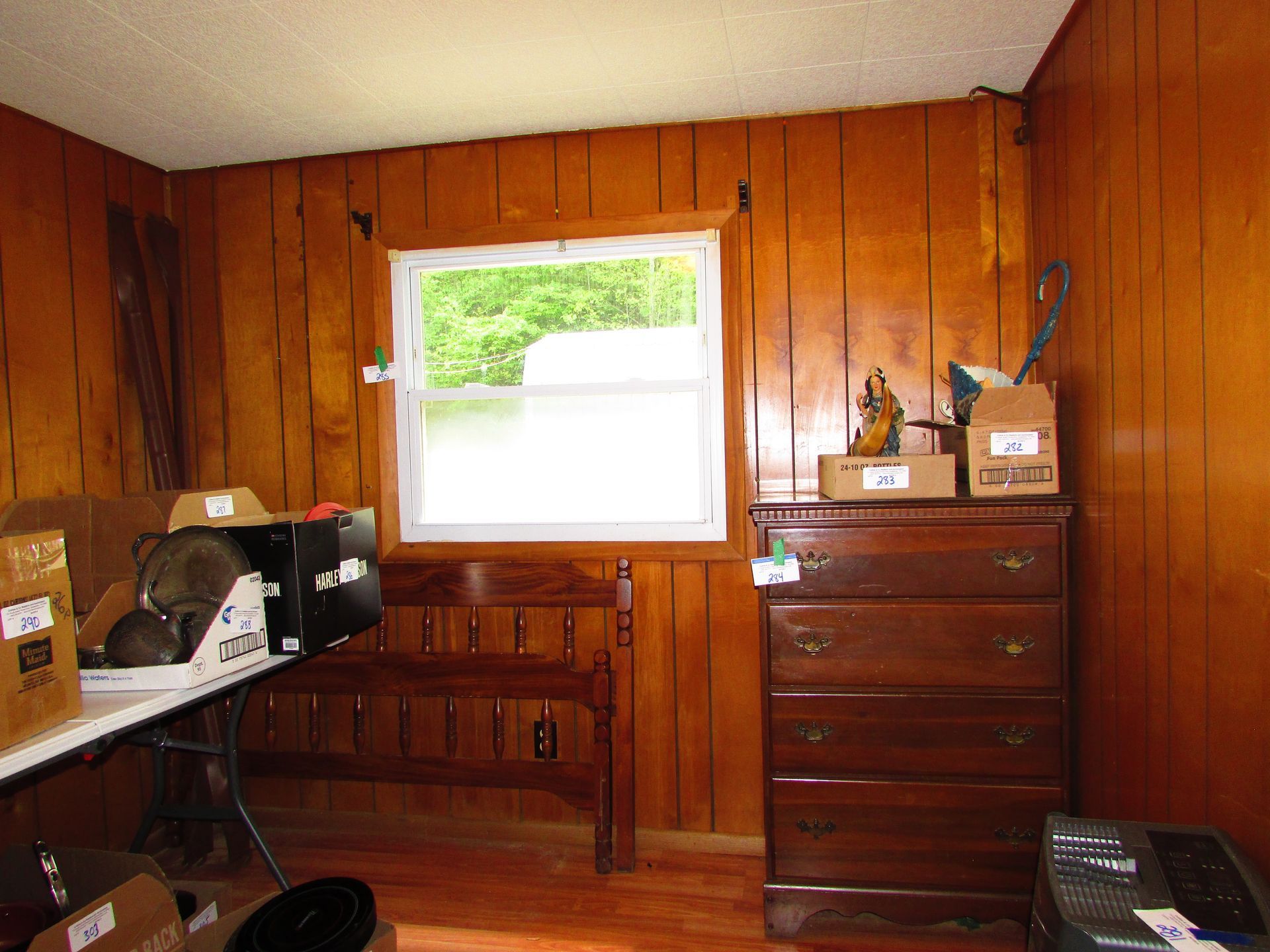 Wood-paneled room with a window, dresser, and cluttered shelves and table.