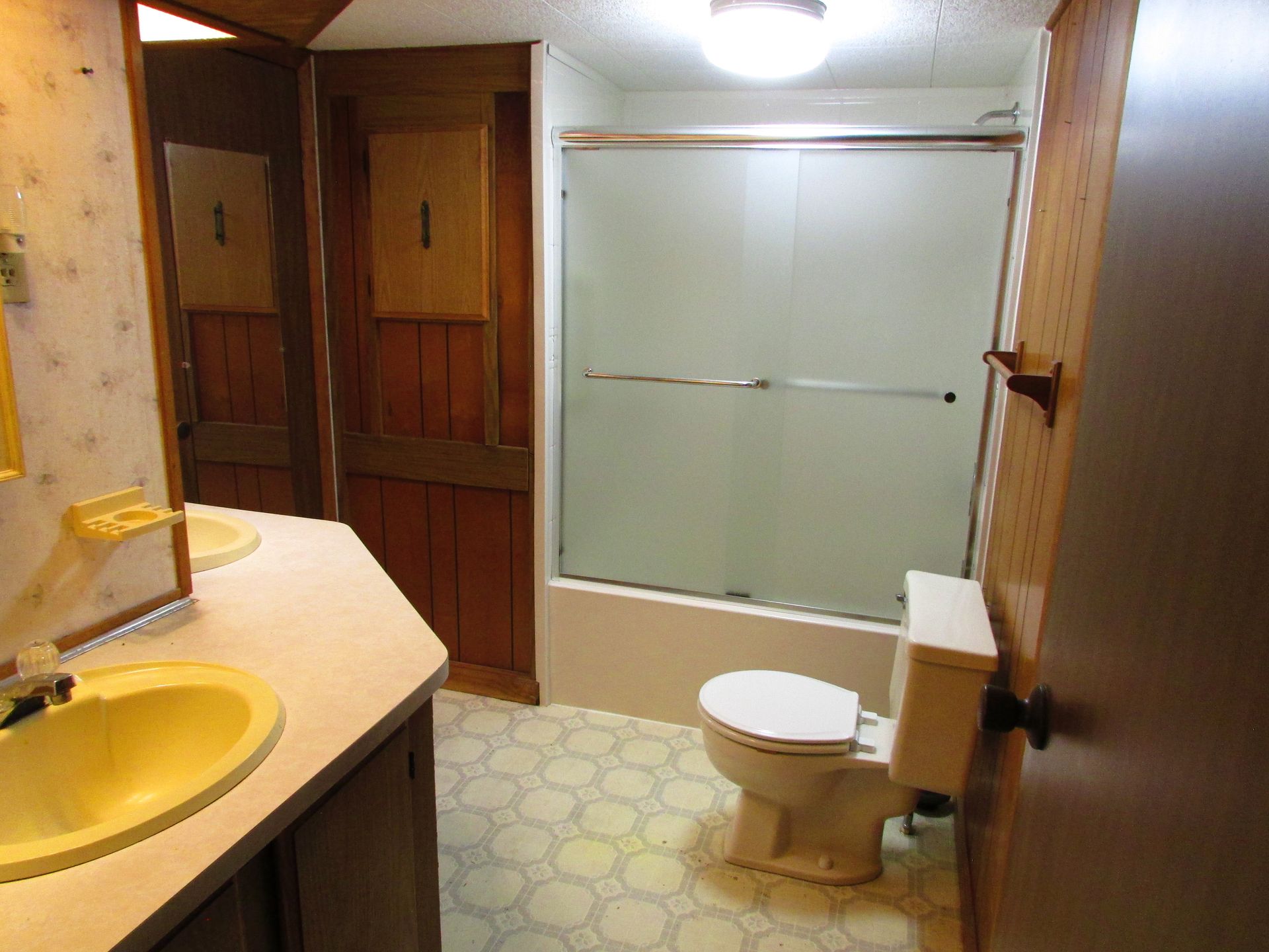 Small bathroom with yellow sink, toilet, frosted glass shower, and wood-paneled walls.