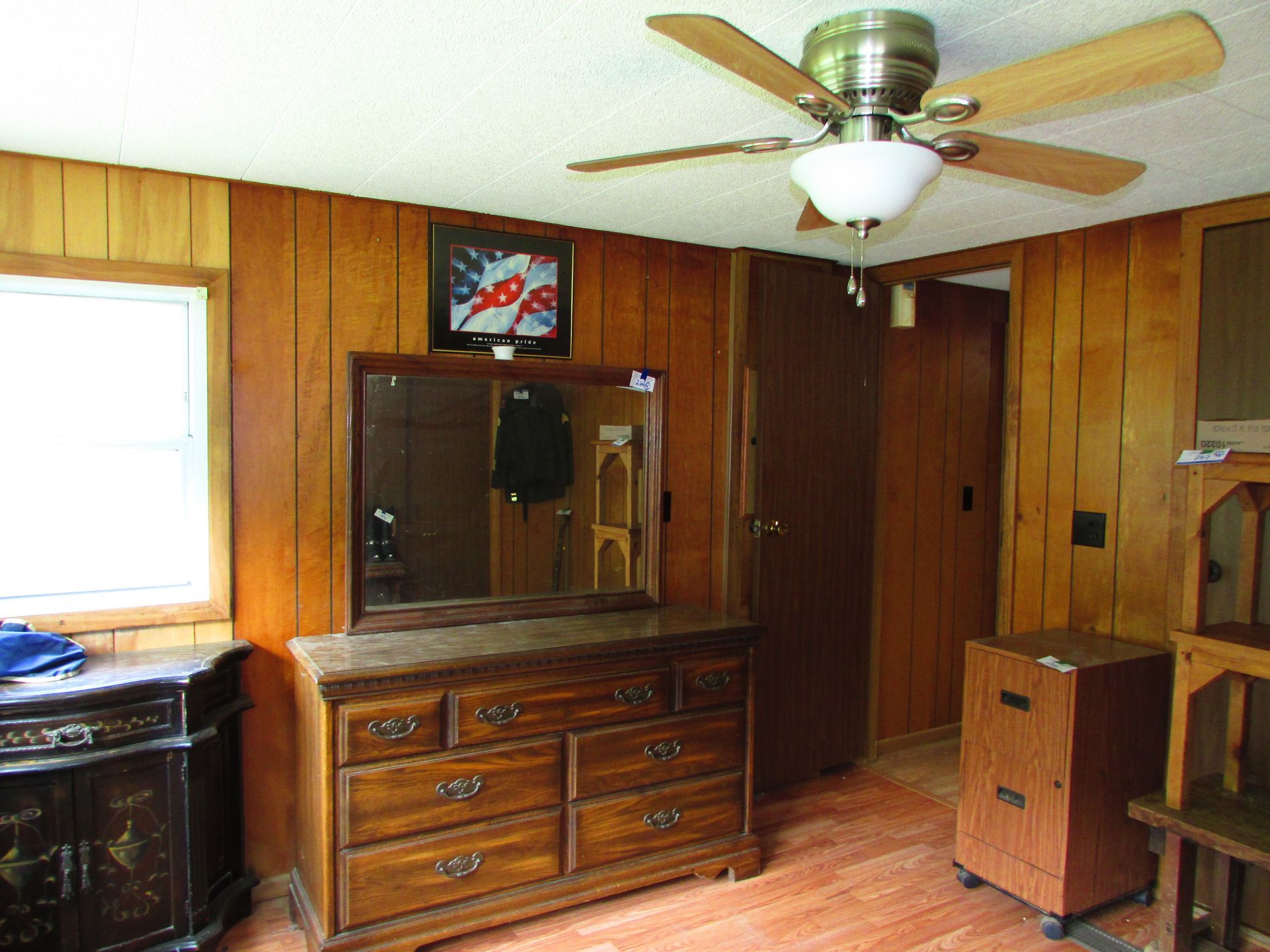 Wood-paneled room with dresser and mirror, ceiling fan, and TV near a window and closed door.