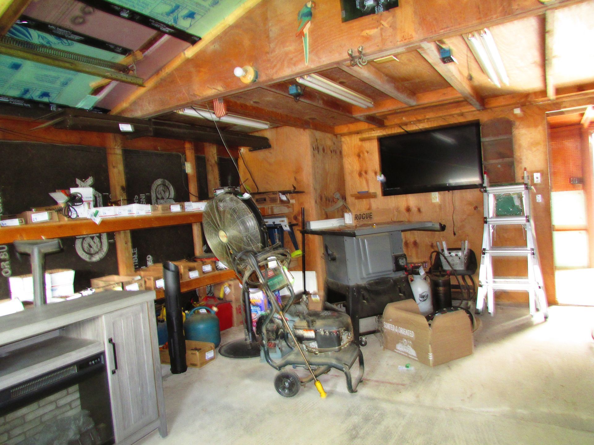 Wood-paneled garage with cluttered shelves, tools, a TV, ladder, and a fan near the center.