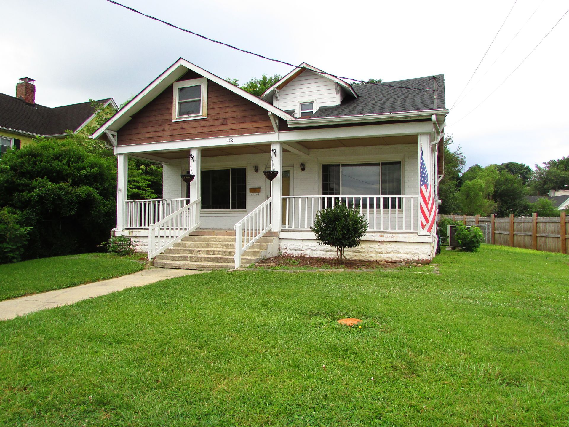 Front view of a small white house with a porch, brick steps, and an American flag on the lawn.
