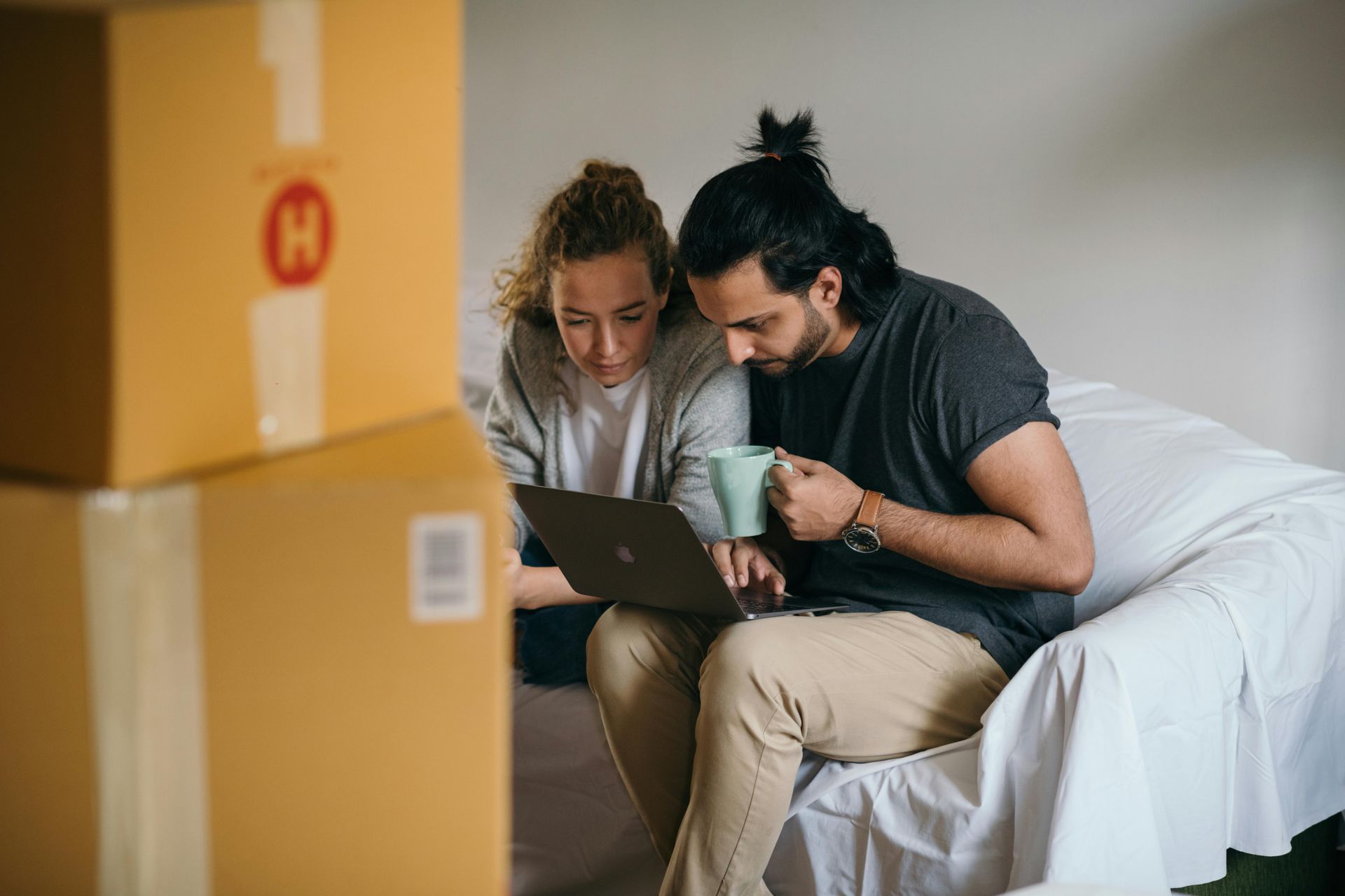 A couple sits together, looking at a laptop. Boxes are nearby. One holds a mug.