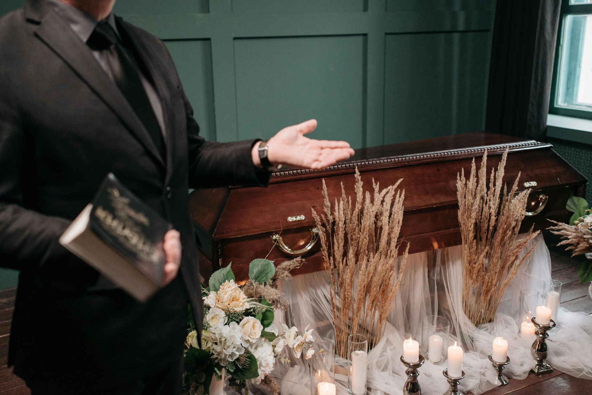 A person in a suit holds a book and gestures toward a wooden casket decorated with dried wheat, candles, and flowers.