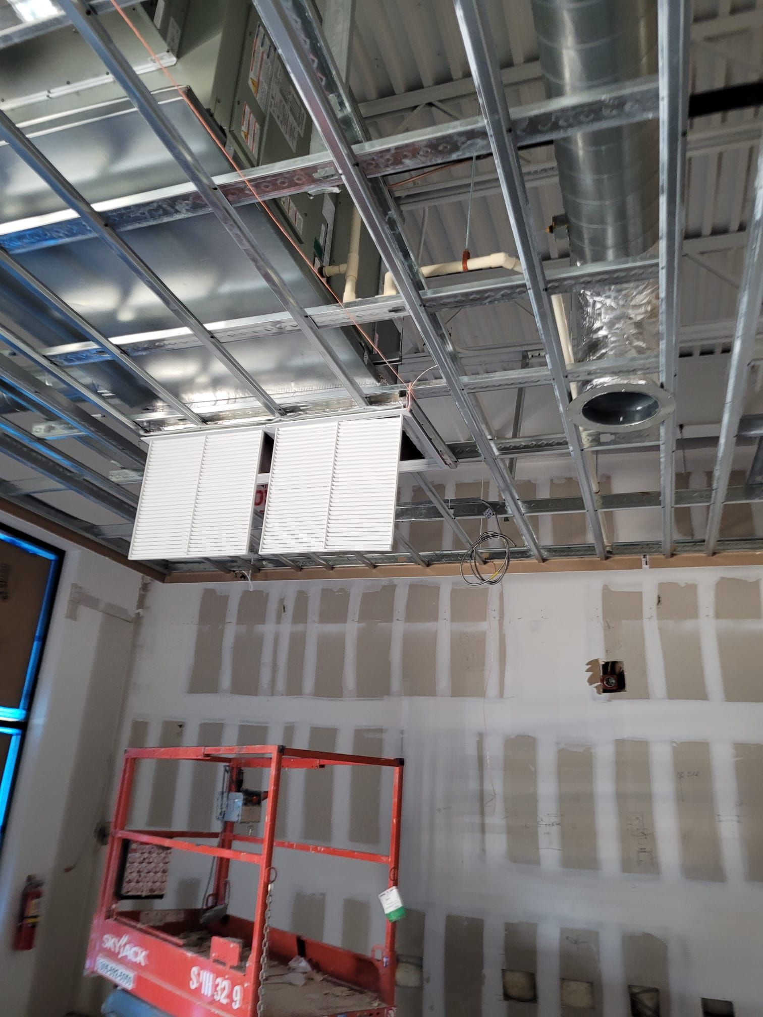 Construction site: ceiling grid with ductwork, white panels, and wall with drywall. Red lift visible.
