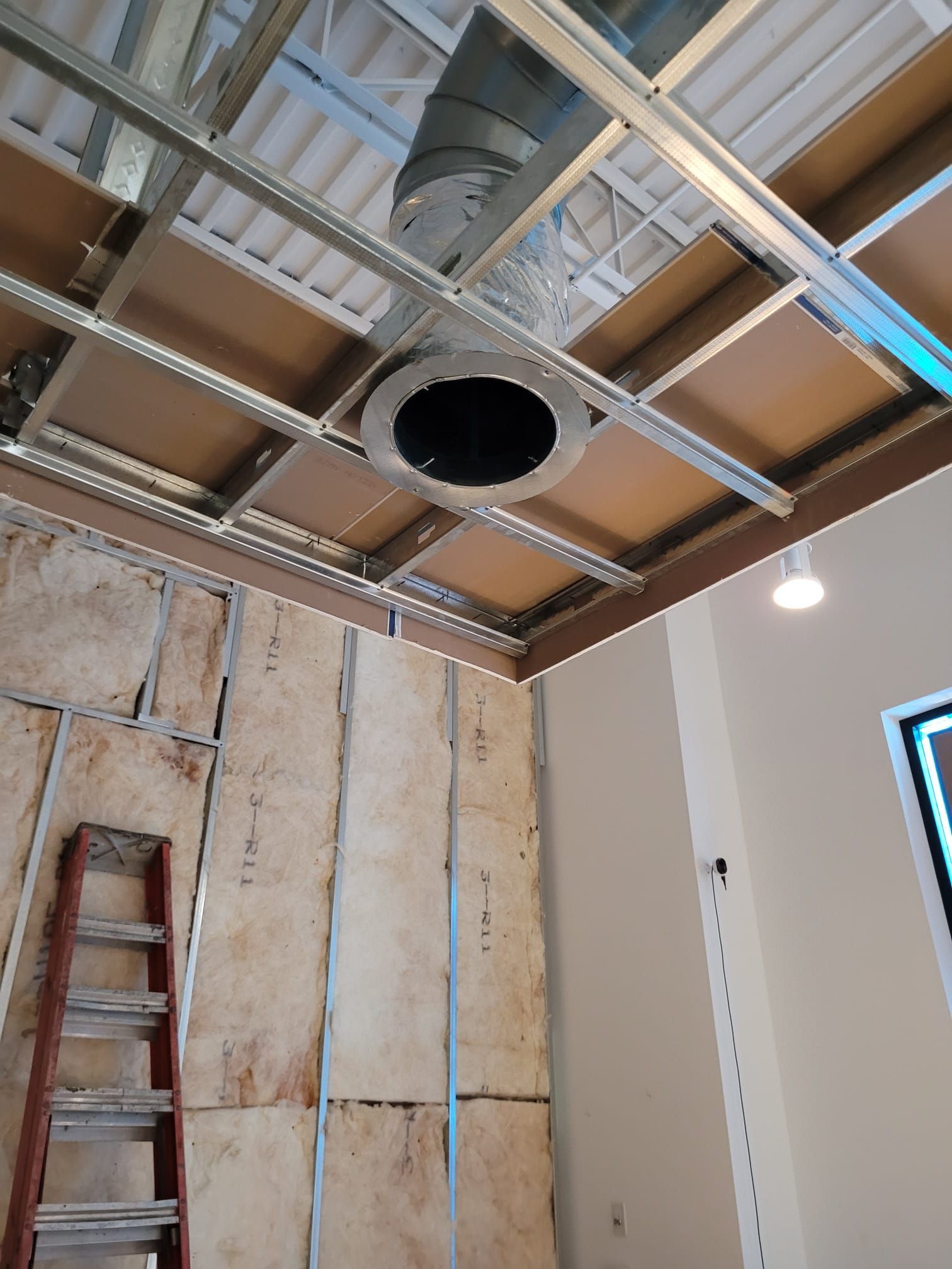 Construction site interior with exposed ceiling framework, ductwork, and insulation. A ladder leans against a partially finished wall.