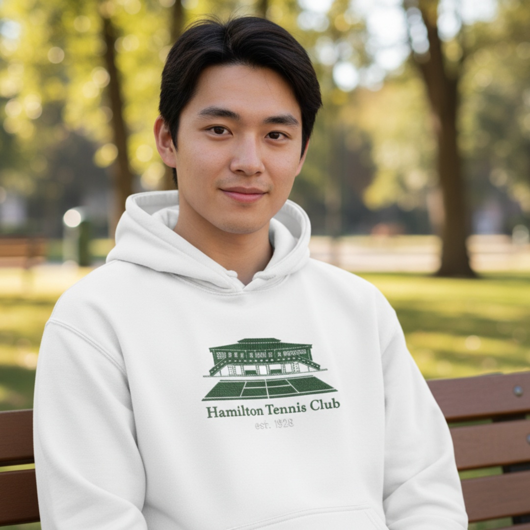 Man in white hoodie with Hamilton Tennis Club logo, smiling outdoors.