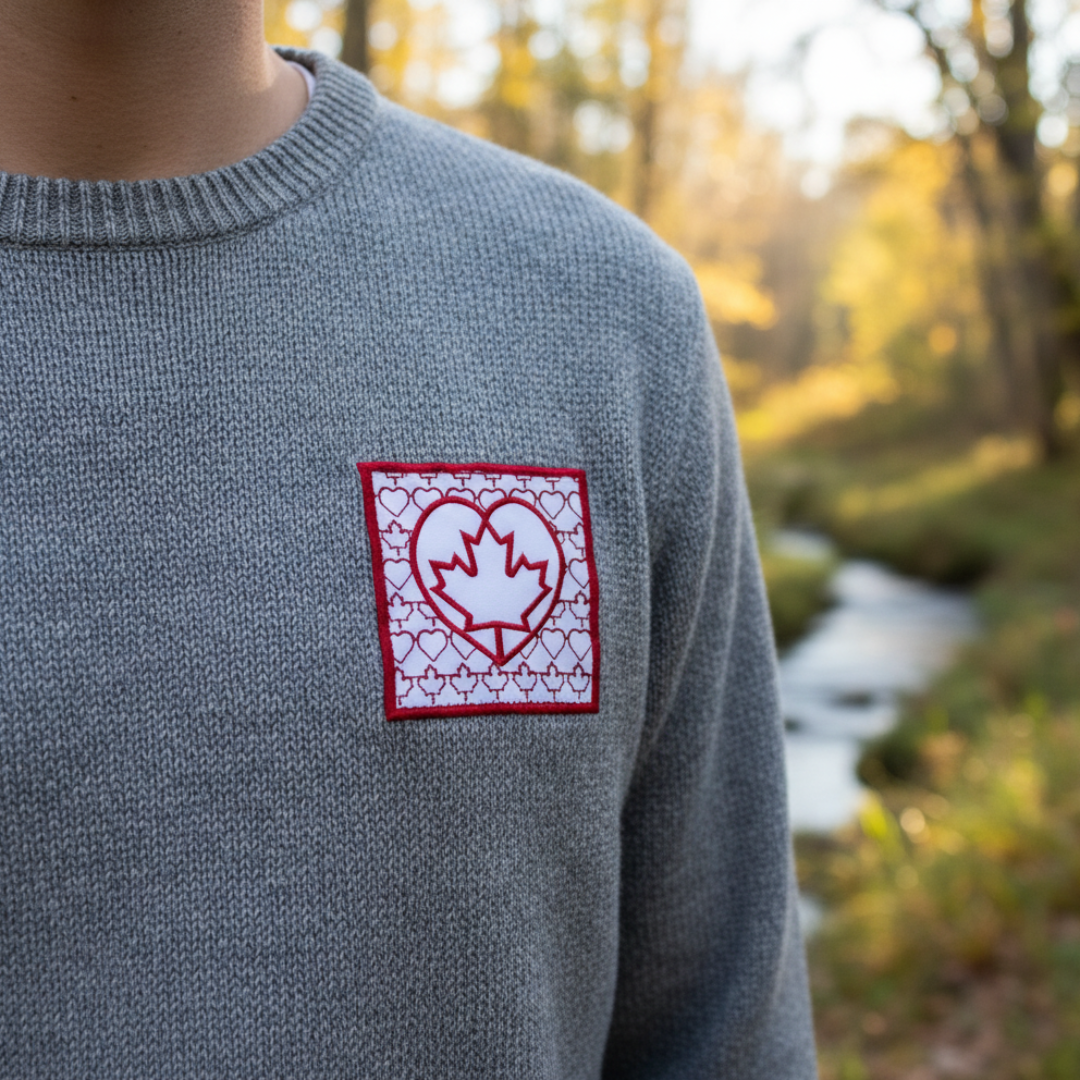 Person in gray sweater with patch: red heart, Canadian maple leaf, and white hearts. Outdoors near a stream.