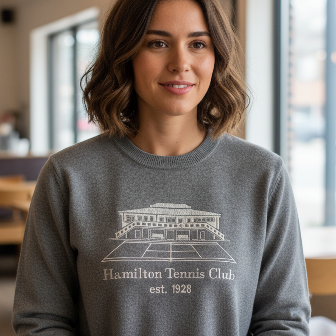 Woman wearing gray Hamilton Tennis Club sweatshirt, smiling indoors.