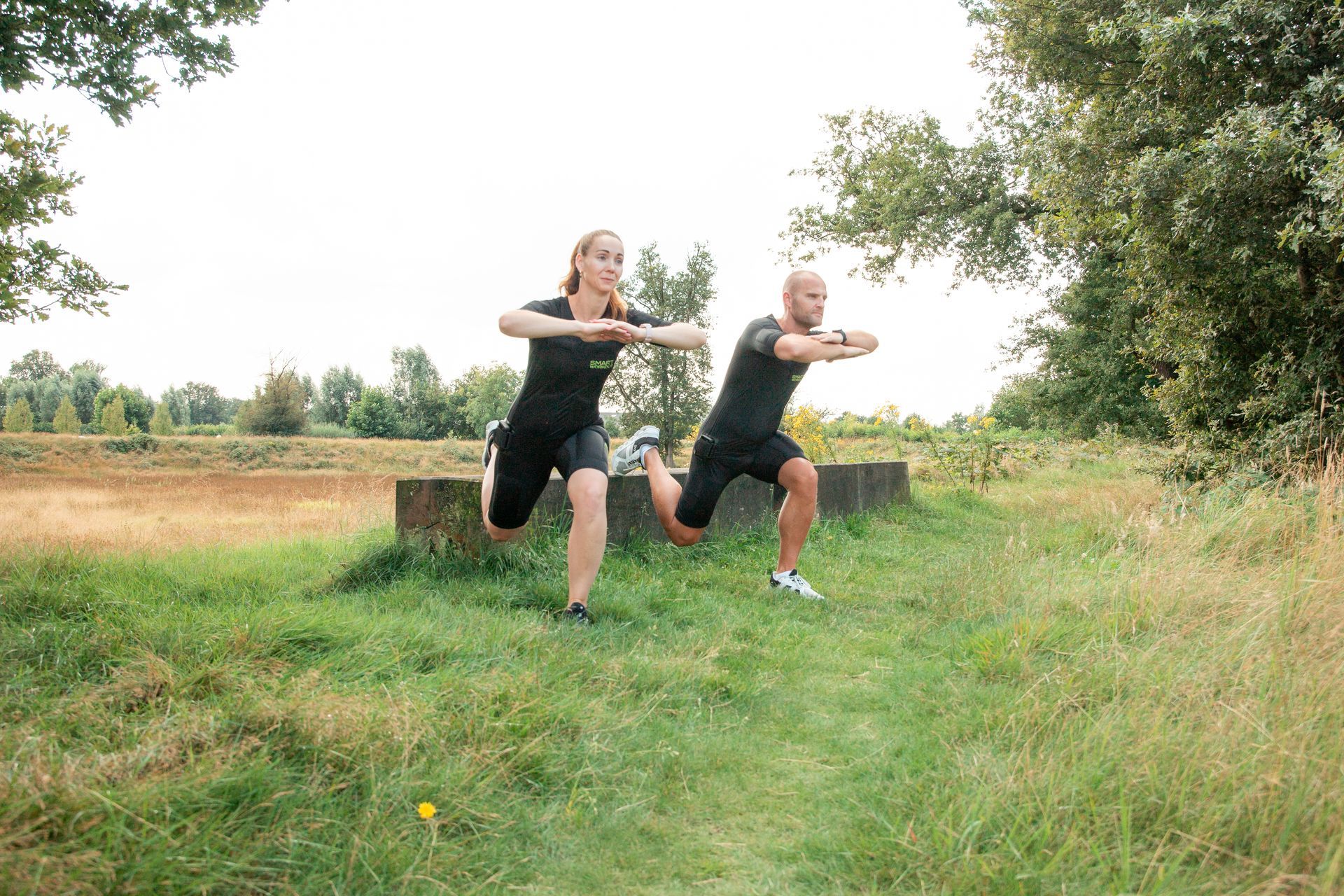 Twee mensen doen lunges op een grasveld vlak bij een betonnen constructie.