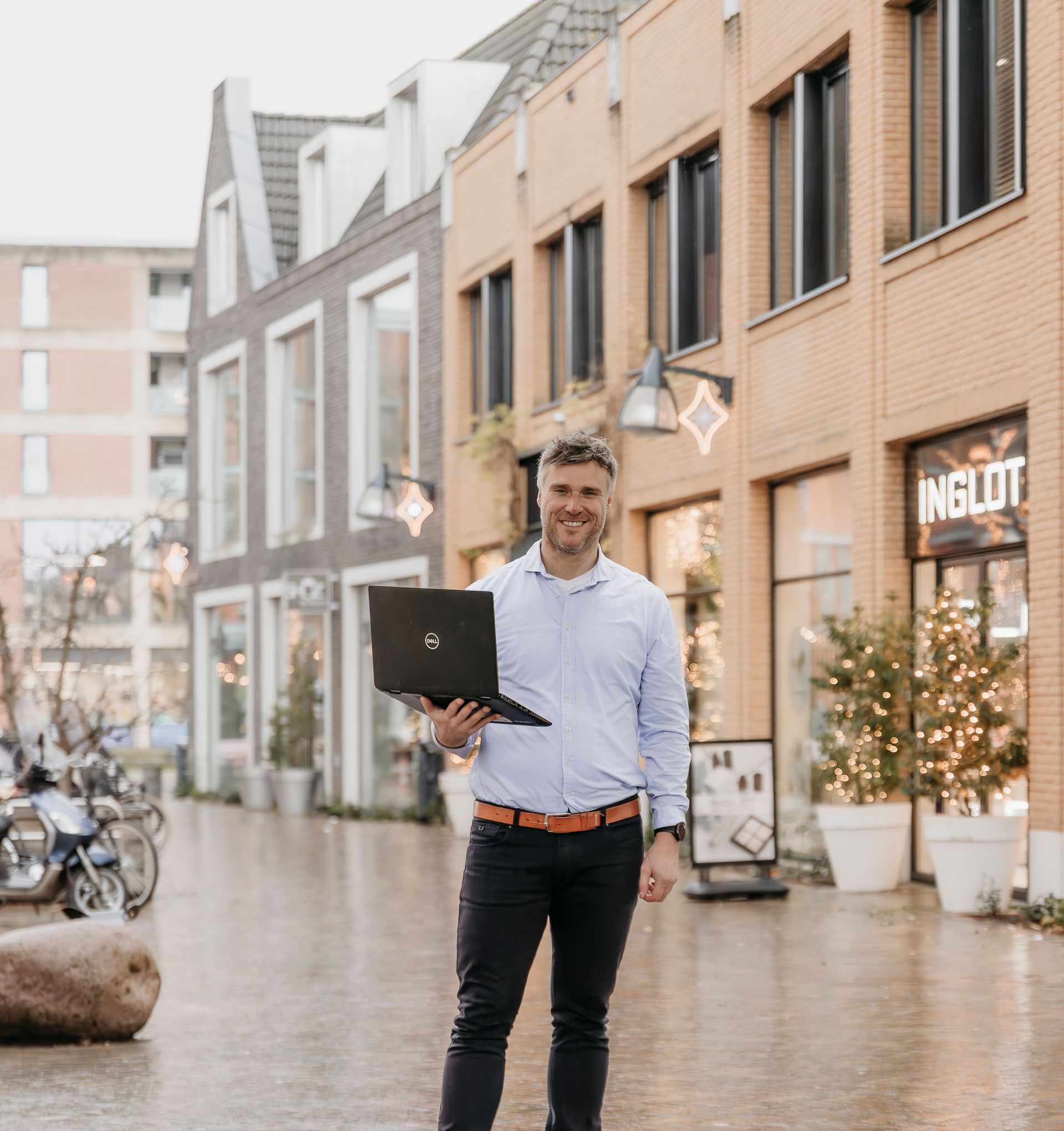 Man in blauw shirt en zwarte broek houdt laptop vast en lacht op straat in de stad, vlak bij winkels.