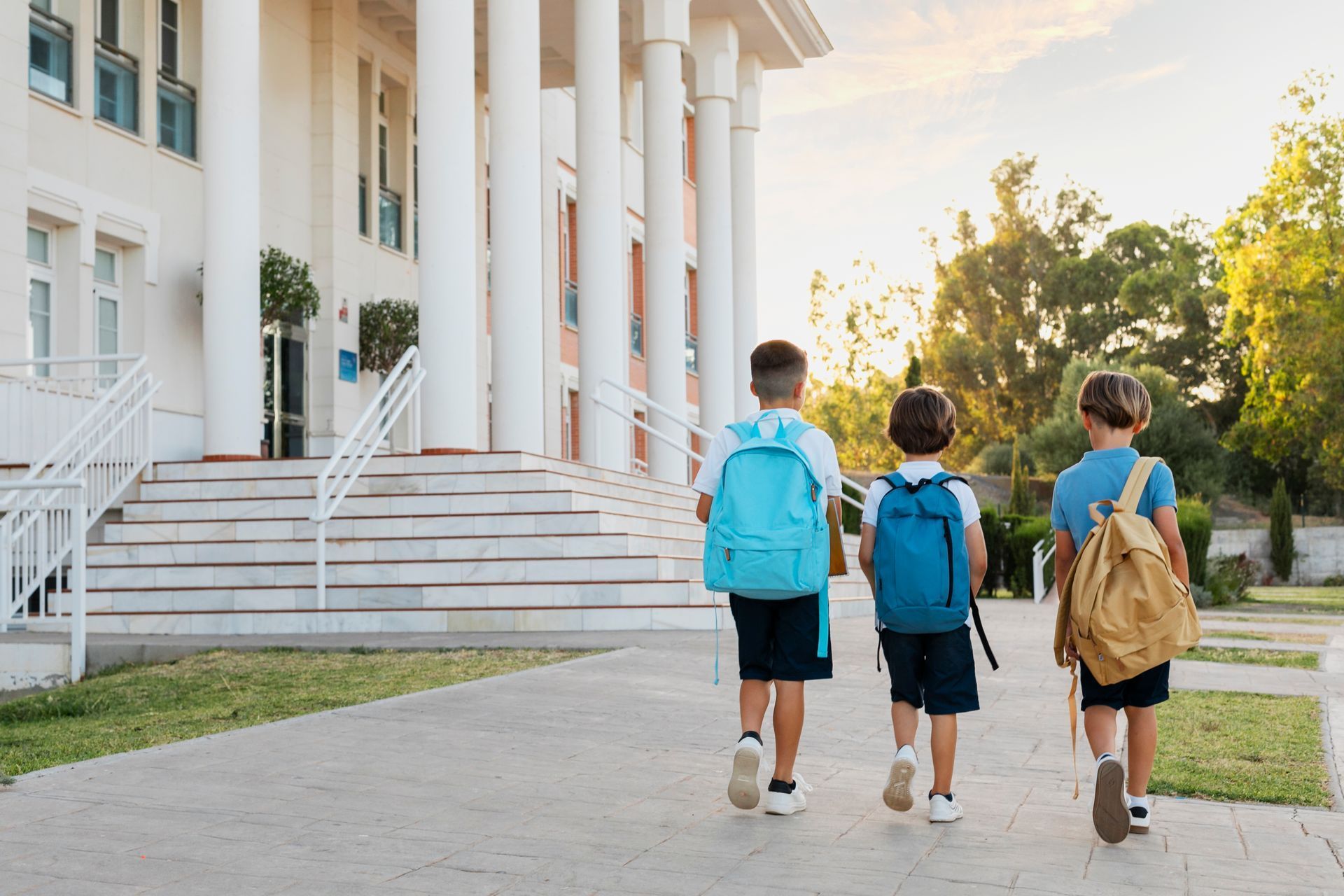 Three children with backpacks walking toward a school building on a sunny day