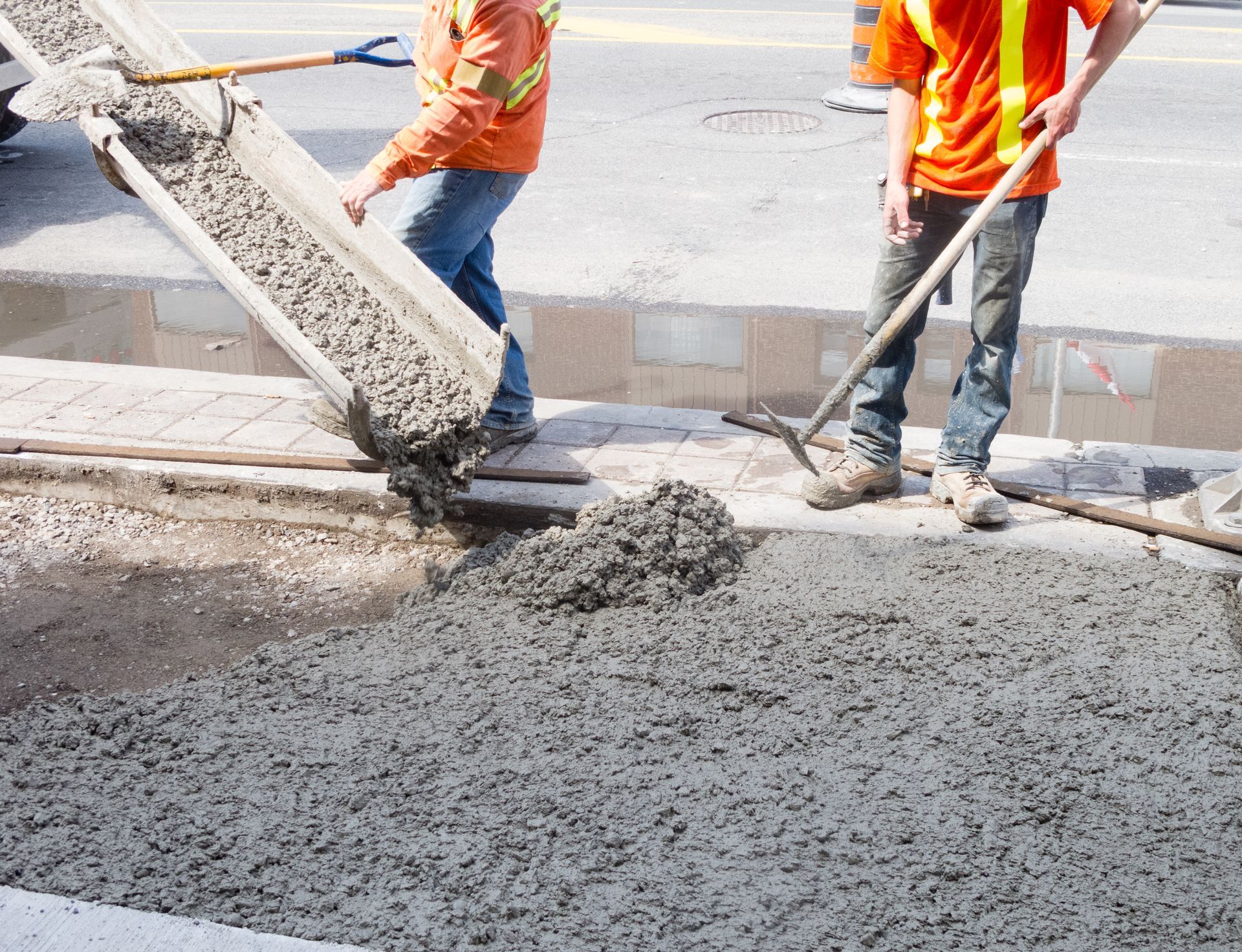 Workers in orange vests pouring wet concrete at a roadside construction site