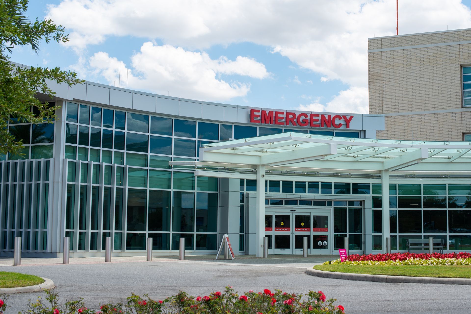 Hospital emergency entrance with glass facade and red āEMERGENCYā sign above a covered walkway.