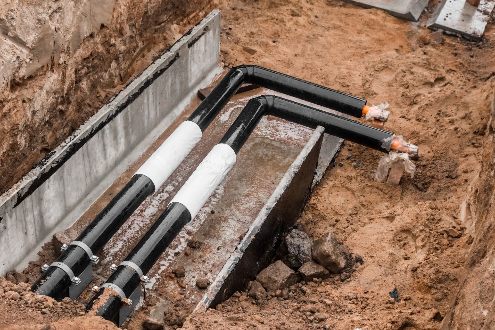 Underground utility pipes and insulated cables installed in an excavated trench beside a concrete wall