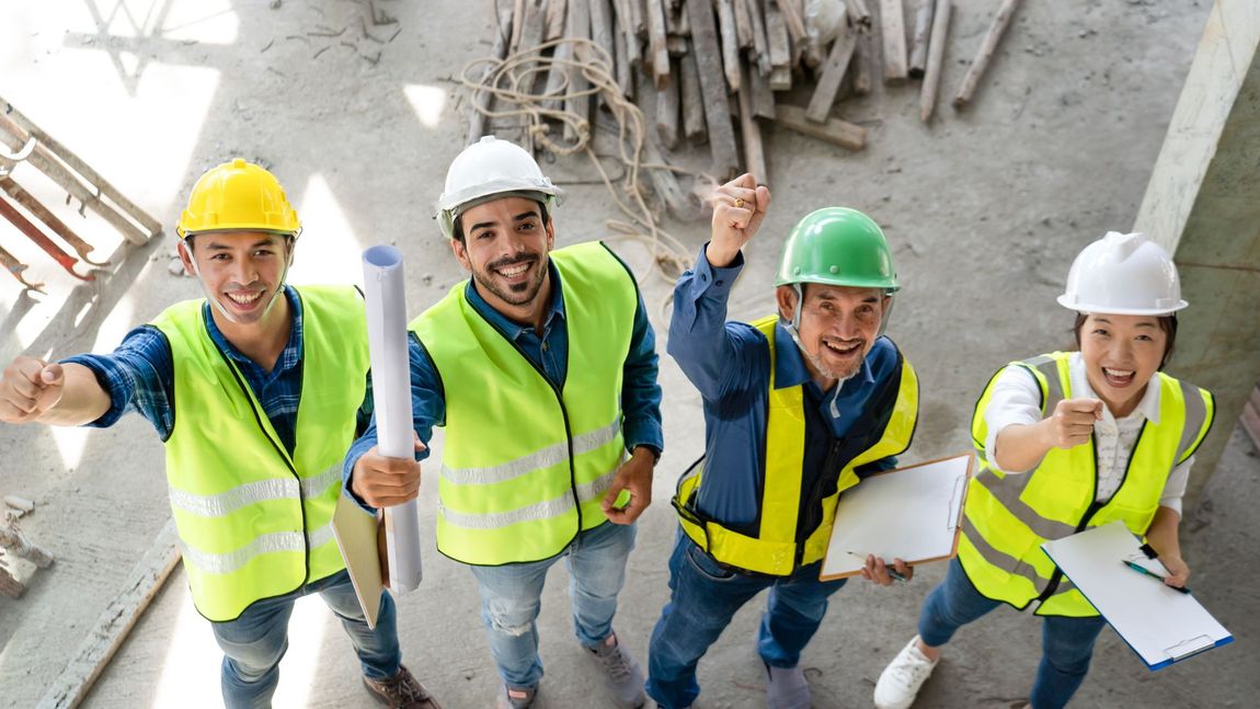 Four construction workers in hard hats and safety vests posing on a building site, smiling and waving up at the camera