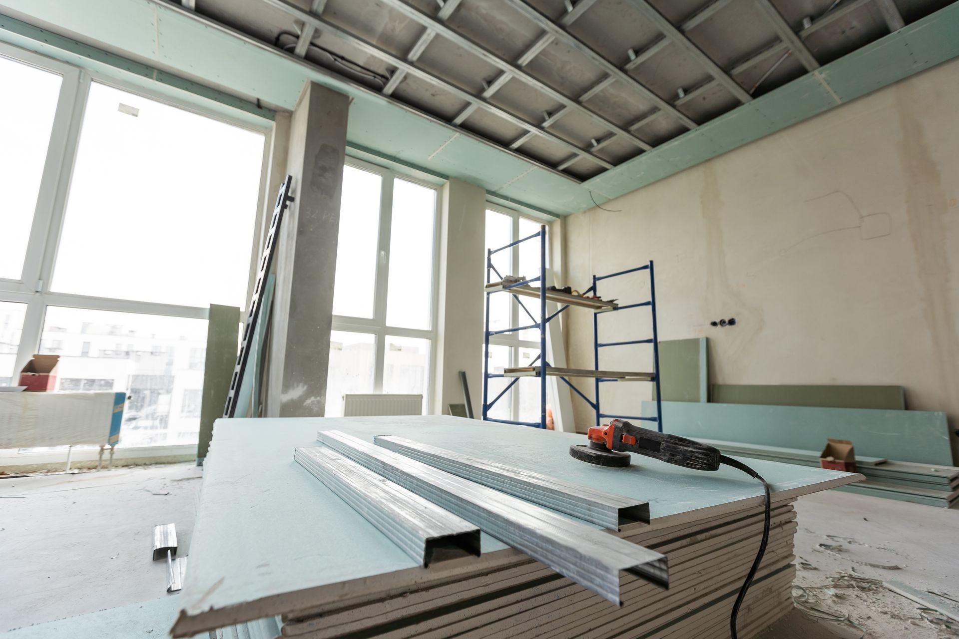 Partially finished room with stacked drywall sheets, a ladder, and construction tools near large windows