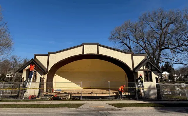 Outdoor band shell stage with cream arch, beige backdrop, and people on a sunny day