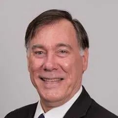 A smiling person wearing a dark suit and white collared shirt against a plain, light gray background.