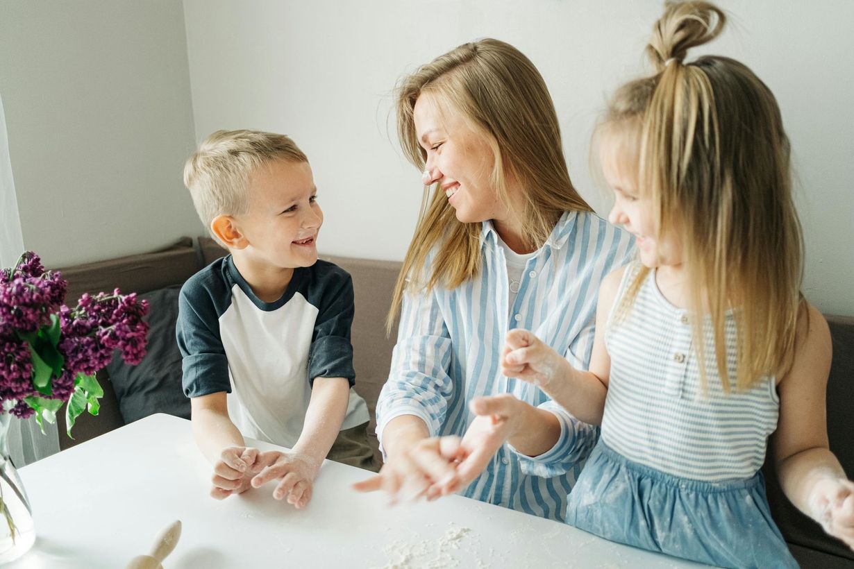 A parent and two children smile while playing with flour on a table next to a bouquet of purple flowers.