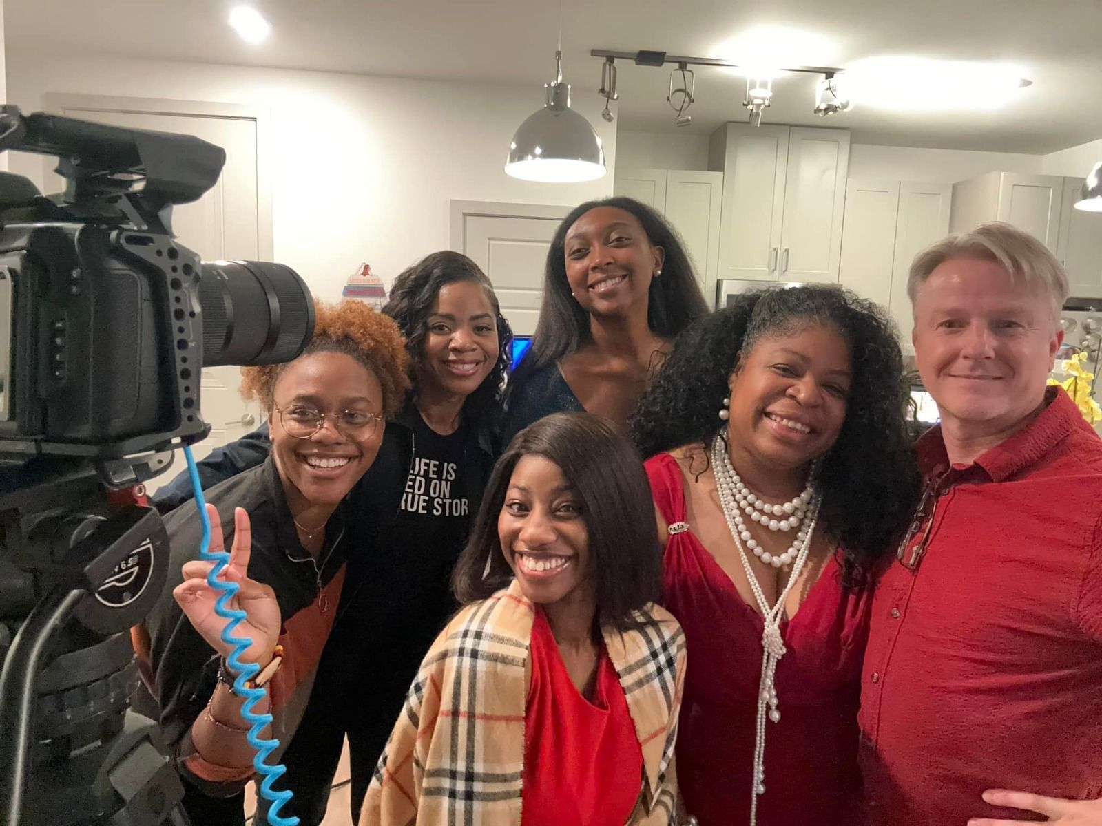 Group of people smiling, posing near a video camera indoors. One person holds up a peace sign.